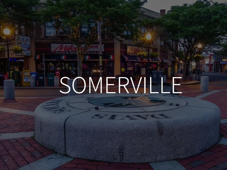 A large stone compass rose with the word 'SOMERVILLE' in the center, located in a town square with storefronts, streetlights, and trees in the background.