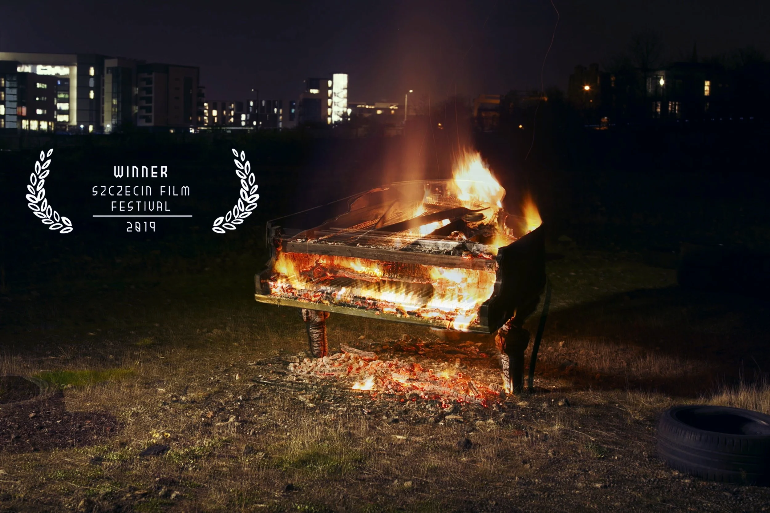 Burning piano on grass with burning embers and flames, documentary editor filmmaker cinematographer. Award overlay reads 'Winner Szczecin Film Festival 2019' on the left side. Instruments in the Architecture: Building the Pianodrome