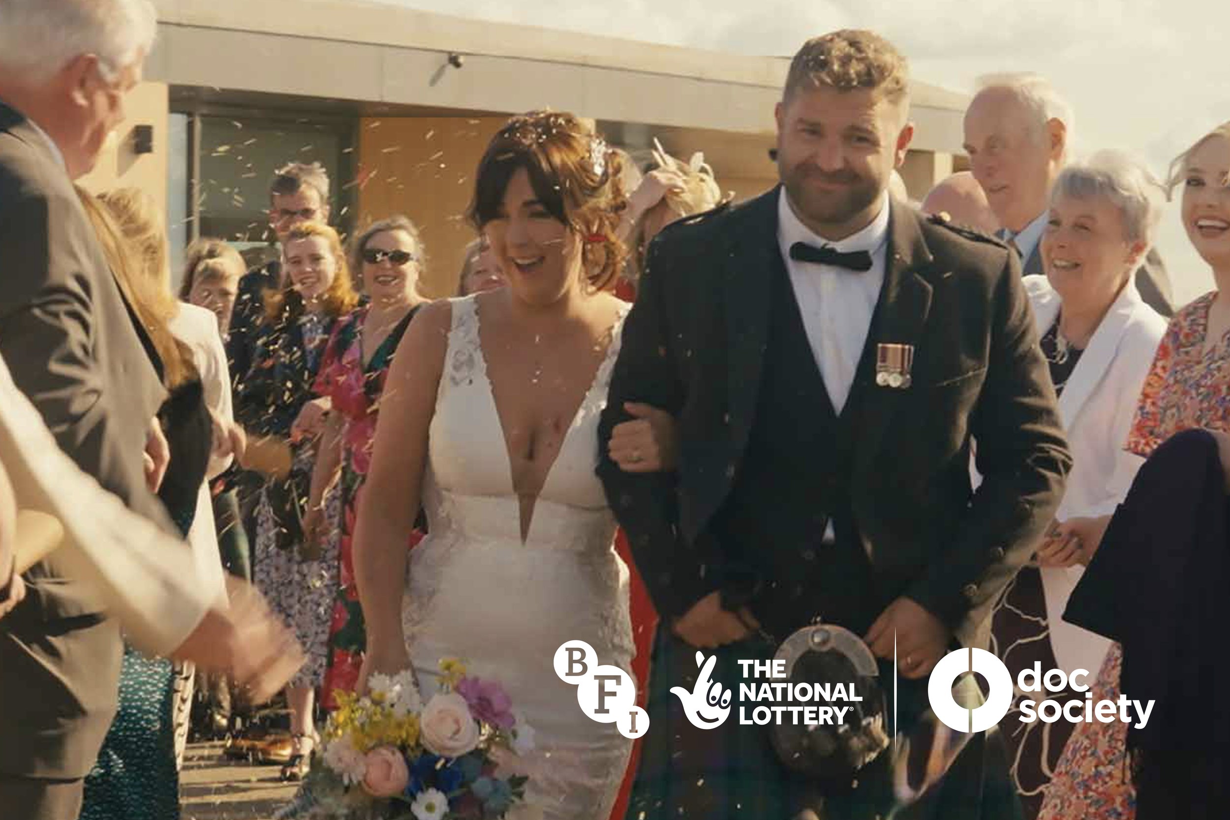 Love & Trouble Documentary Film Amy Hardie Will Hewitt Editor. A newlywed couple, with the bride holding a bouquet, walking arm-in-arm through a crowd of smiling people outdoors, celebrating their wedding.