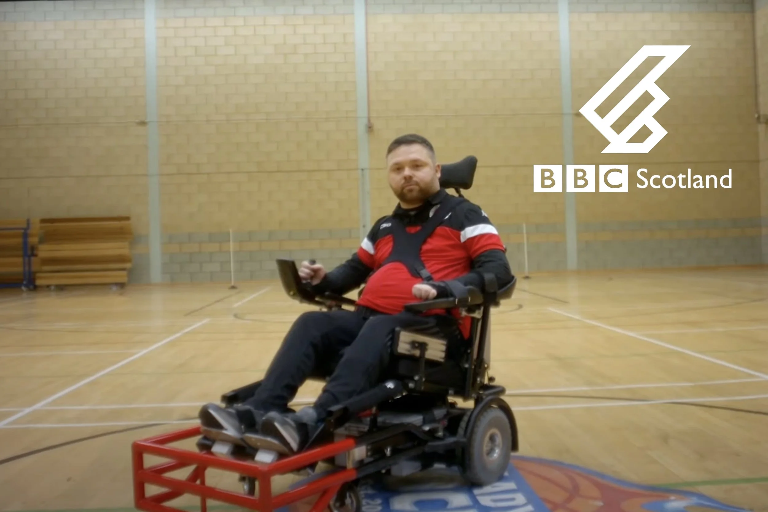 A man in a red and black sports jersey sitting in a motorized wheelchair in an indoor gymnasium, with the BBC Scotland logo overlayed on the right side. Playing Powerchair Football documentary director, filmmaker