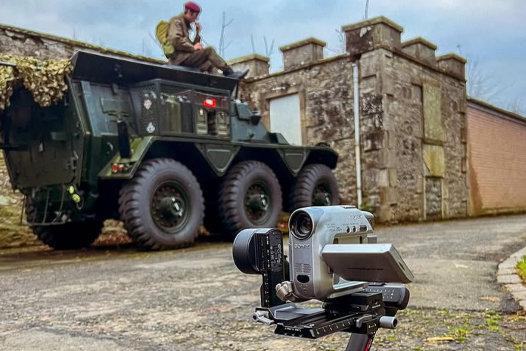 A vintage film camera on a tripod capturing a scene with a military-style vehicle and a person sitting on top of it against a stone wall. filmmaker cinematographer documentary editor Dougie & me