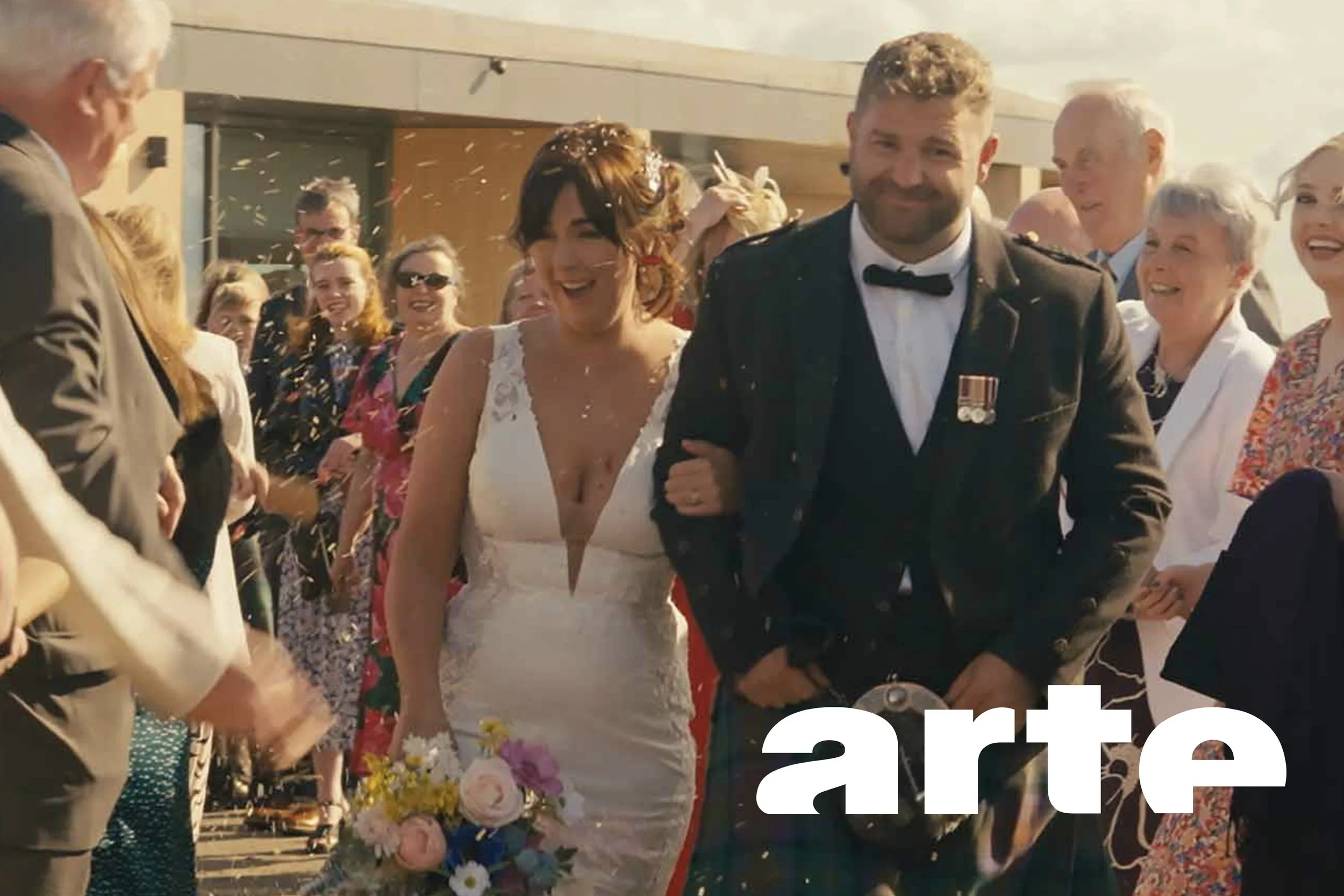 A bride and groom walking arm in arm, surrounded by smiling wedding guests, with confetti in the air during a celebration. Love & Trouble Documentary Editor, Arte TV filmmaker cinematographer