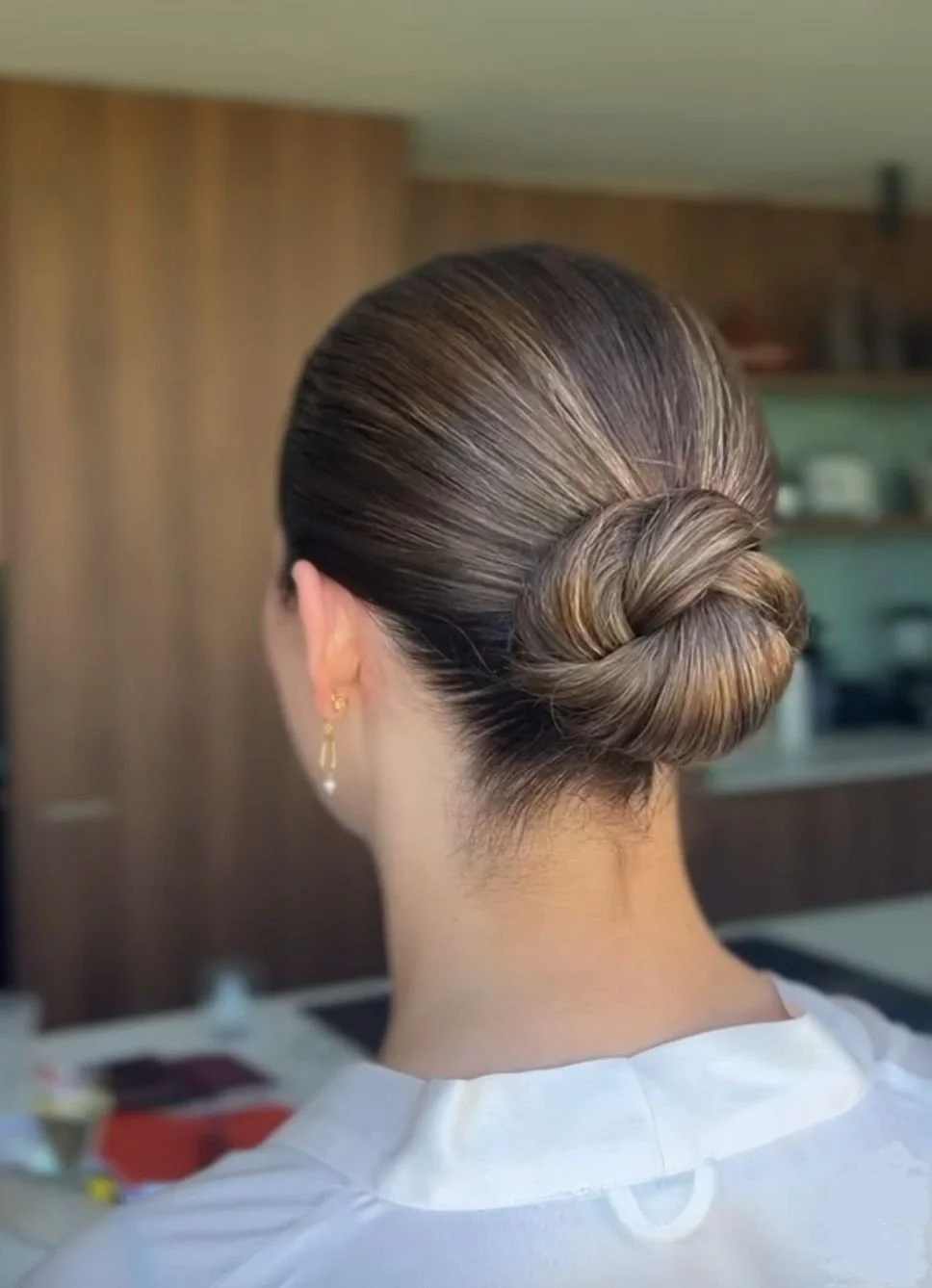 Back view of a woman with her hair styled in a neatly twisted and pinned bun, wearing a white top and gold earrings.