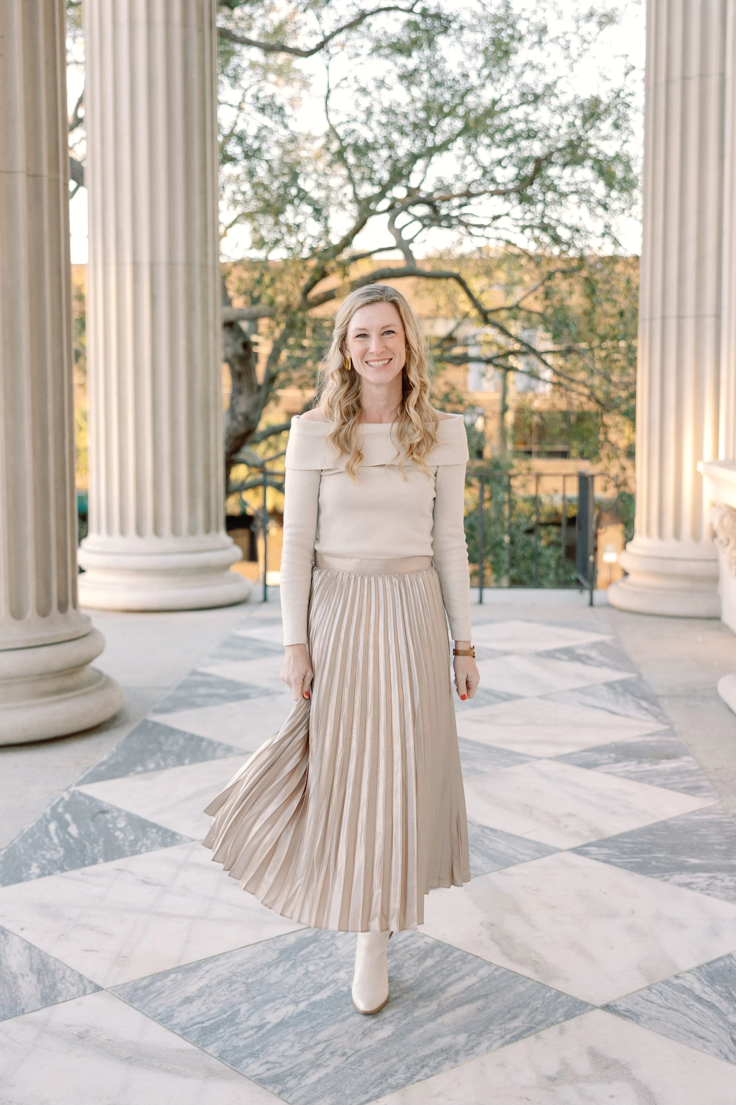 A woman with long blonde hair wearing a cream-colored off-the-shoulder top and a pleated metallic silver skirt, standing between large white columns in an outdoor setting.
