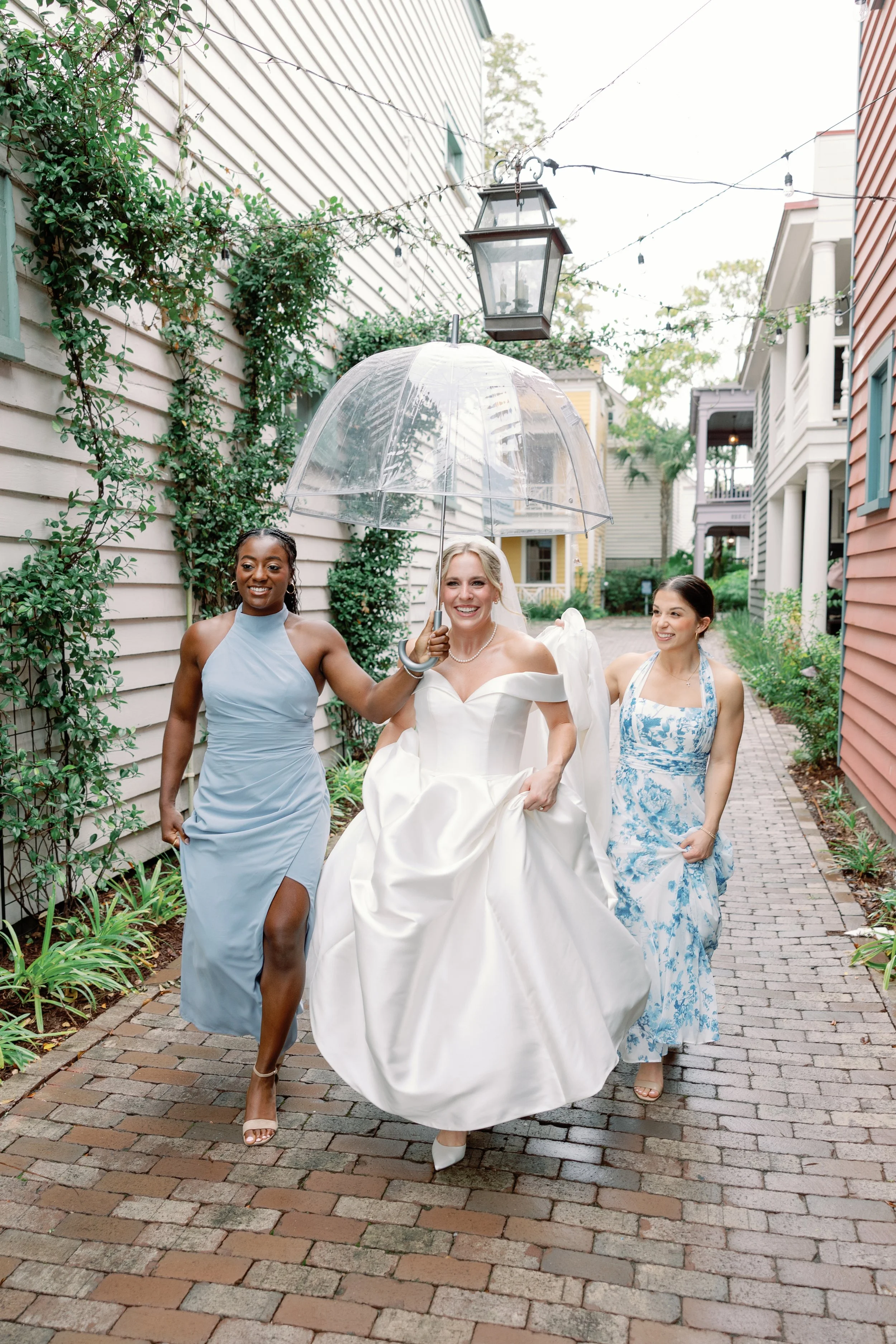 Bride in a white wedding gown, smiling, walking with two women, one in a light blue dress and the other in a blue and white floral dress, while holding a clear umbrella. They are walking along a brick pathway with greenery and colorful houses in the background on a cloudy day.