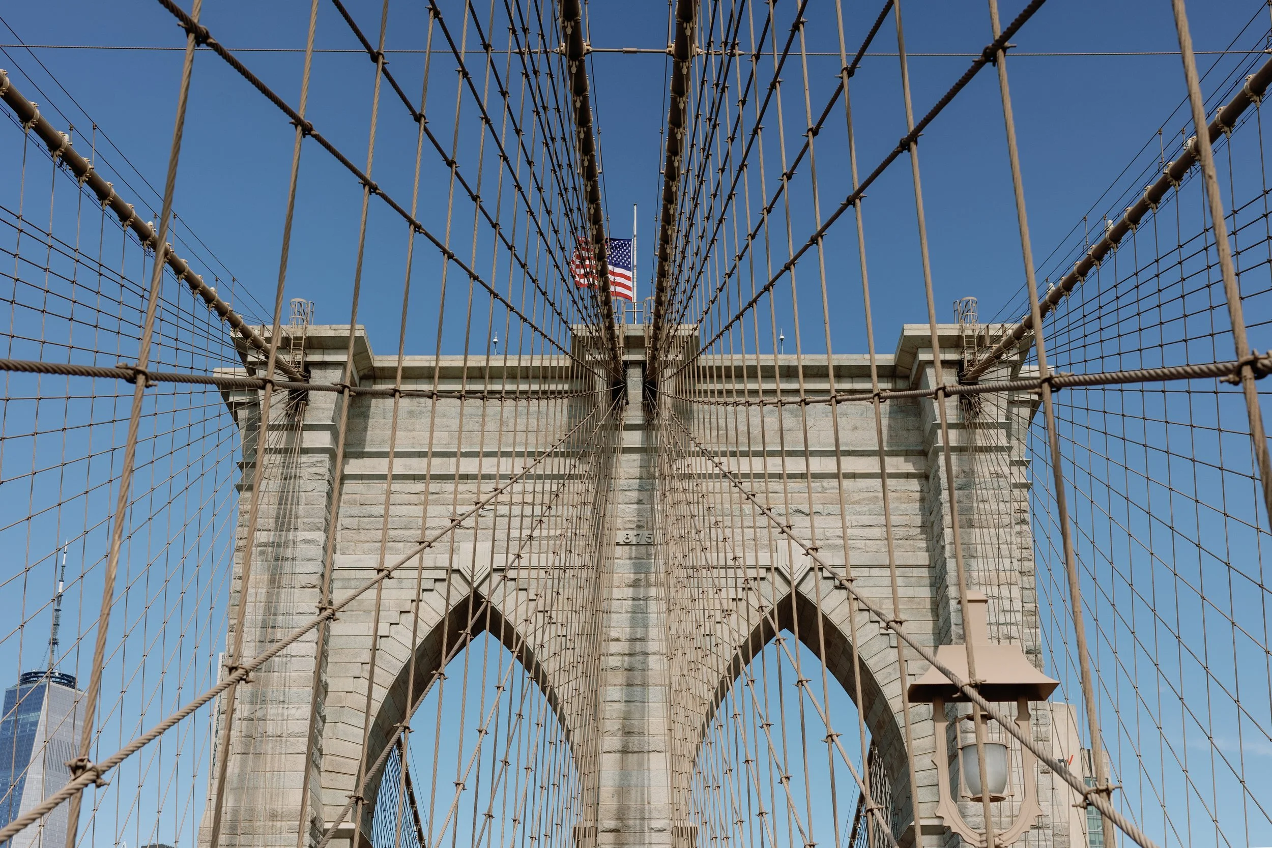 View of the Brooklyn Bridge from below, showing the stone towers, bridge cables, and American flags against a blue sky.