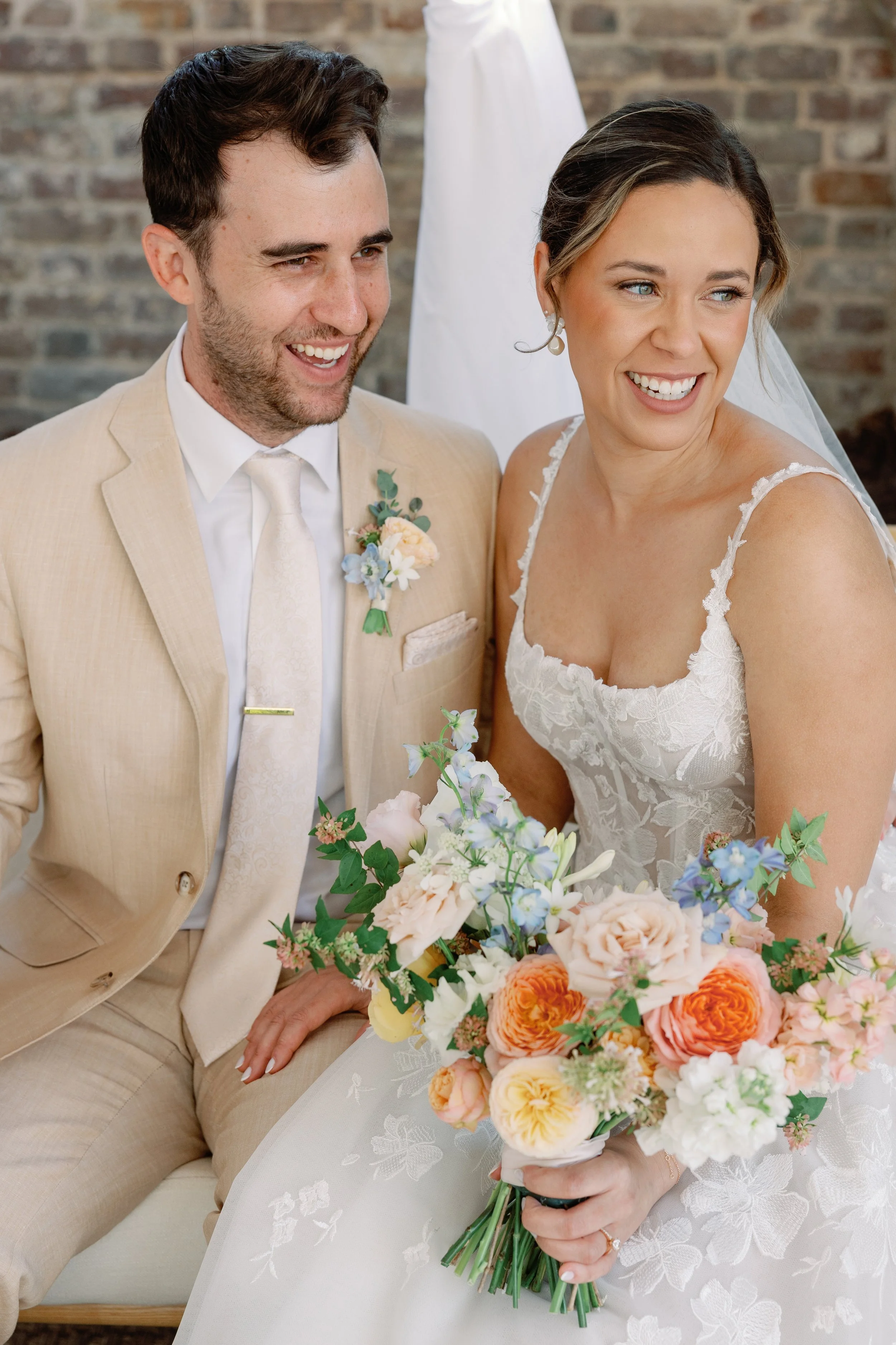 A newly married couple sitting together at their wedding, smiling. The bride is holding a bouquet of colorful flowers while wearing a white lace wedding dress, and the groom is wearing a cream-colored suit with a boutonnière.