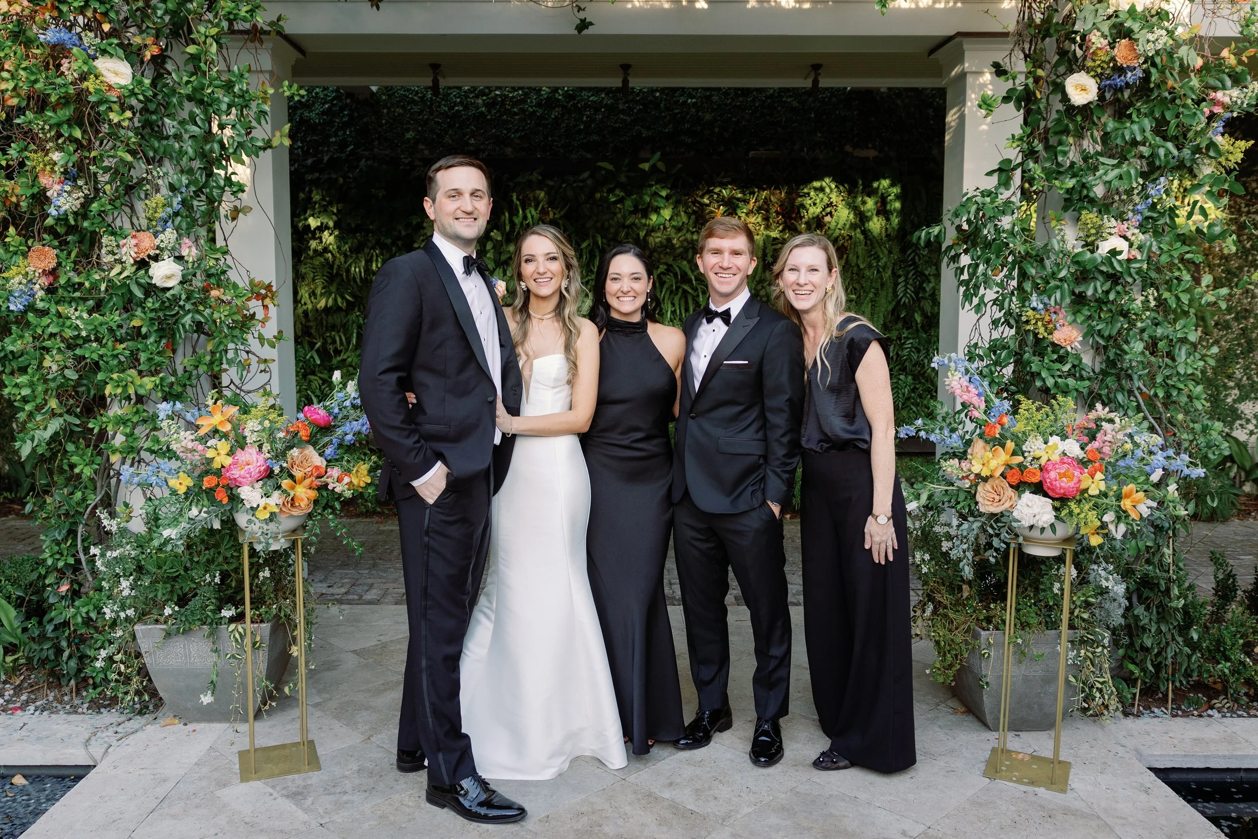 Group of five people dressed in formal attire, smiling, standing in front of a floral arch at a wedding or special event.