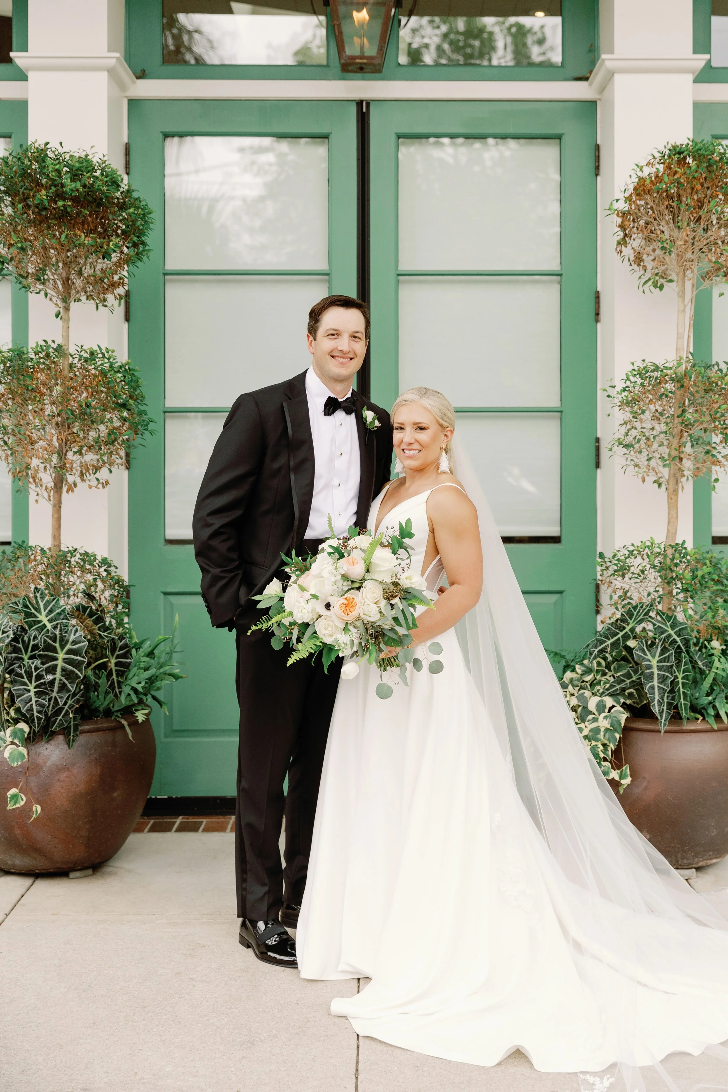 A newlywed couple posing for a wedding photo in front of a green door with large potted plants on either side. The groom is wearing a black tuxedo with a bow tie, and the bride is in a white wedding gown holding a bouquet of flowers.
