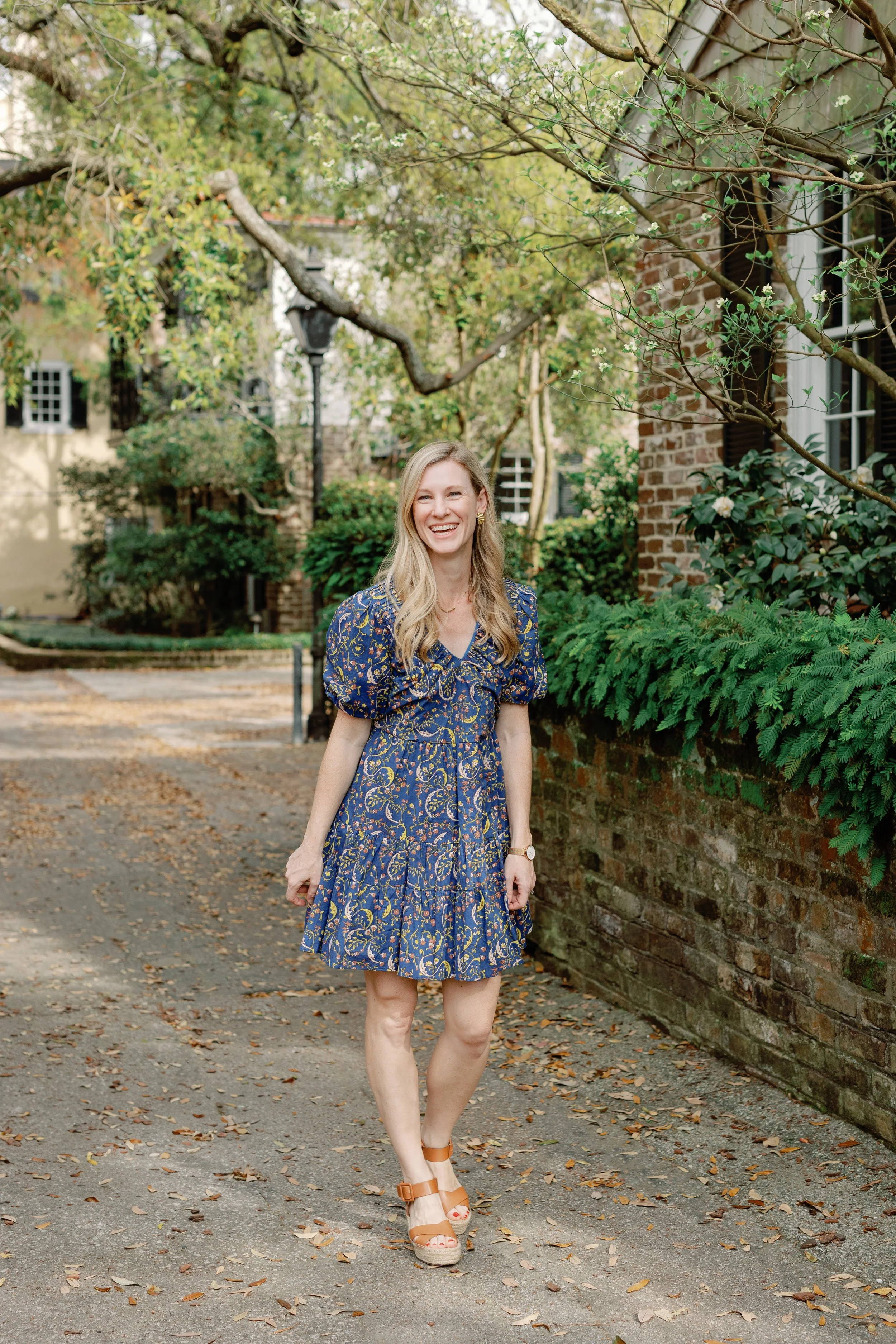 A woman with blonde hair, wearing a navy blue dress with a floral pattern and brown sandals, standing outdoors on a leaf-covered pathway, smiling at the camera with a background of green trees, shrubs, a brick wall, and residential buildings.