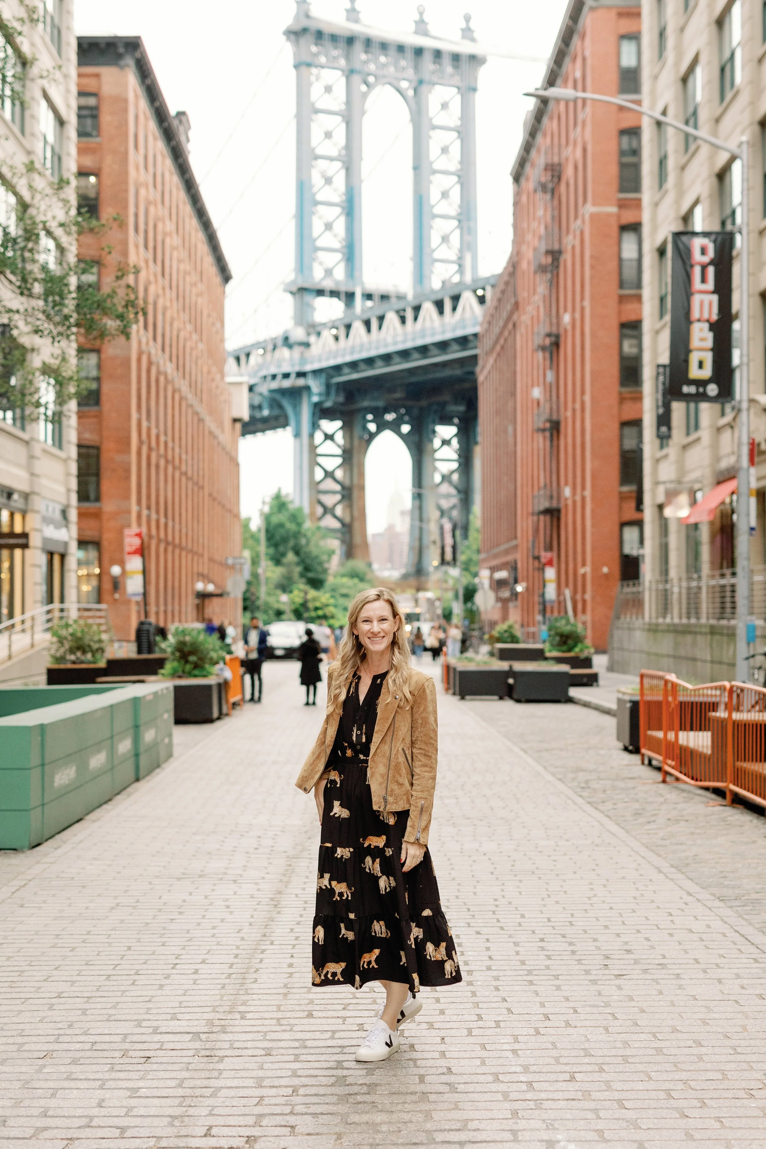 A woman smiling and walking on a cobblestone street with the Manhattan Bridge in the background. She is wearing a black dress with tiger prints, a brown leather jacket, and white sneakers. The street is lined with red brick buildings and some pedestrians are visible in the distance.
