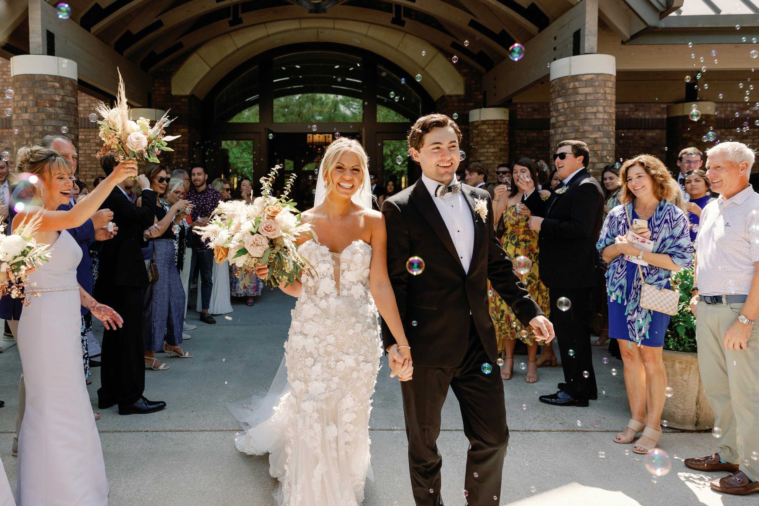 A newlywed couple walking hand in hand out of a building, surrounded by friends and family celebrating with bubbles on a sunny day.