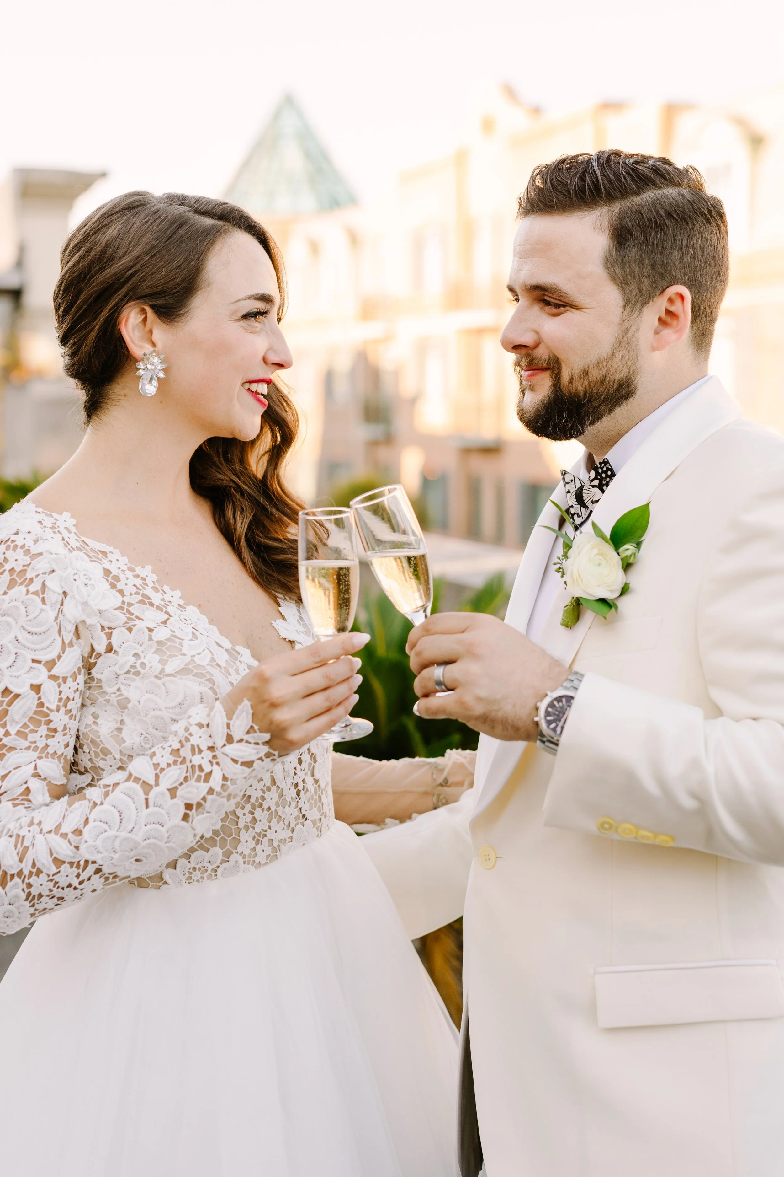 A bride and groom in wedding attire celebrating, raising champagne glasses and gazing into each other's eyes outdoors with a building in the background.