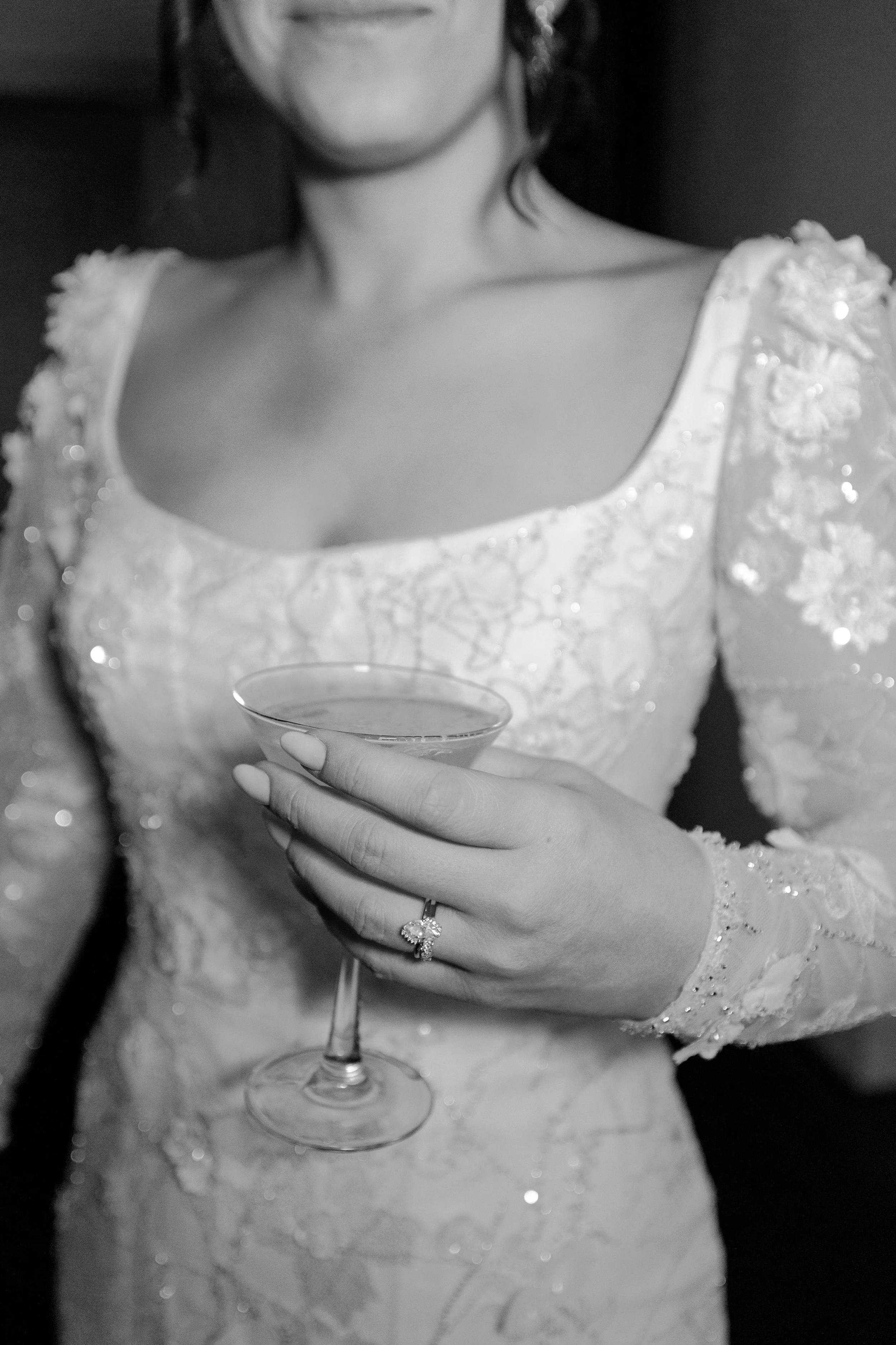 A woman in an elegant, embellished dress is holding a cocktail glass in her right hand, wearing a ring on her finger.