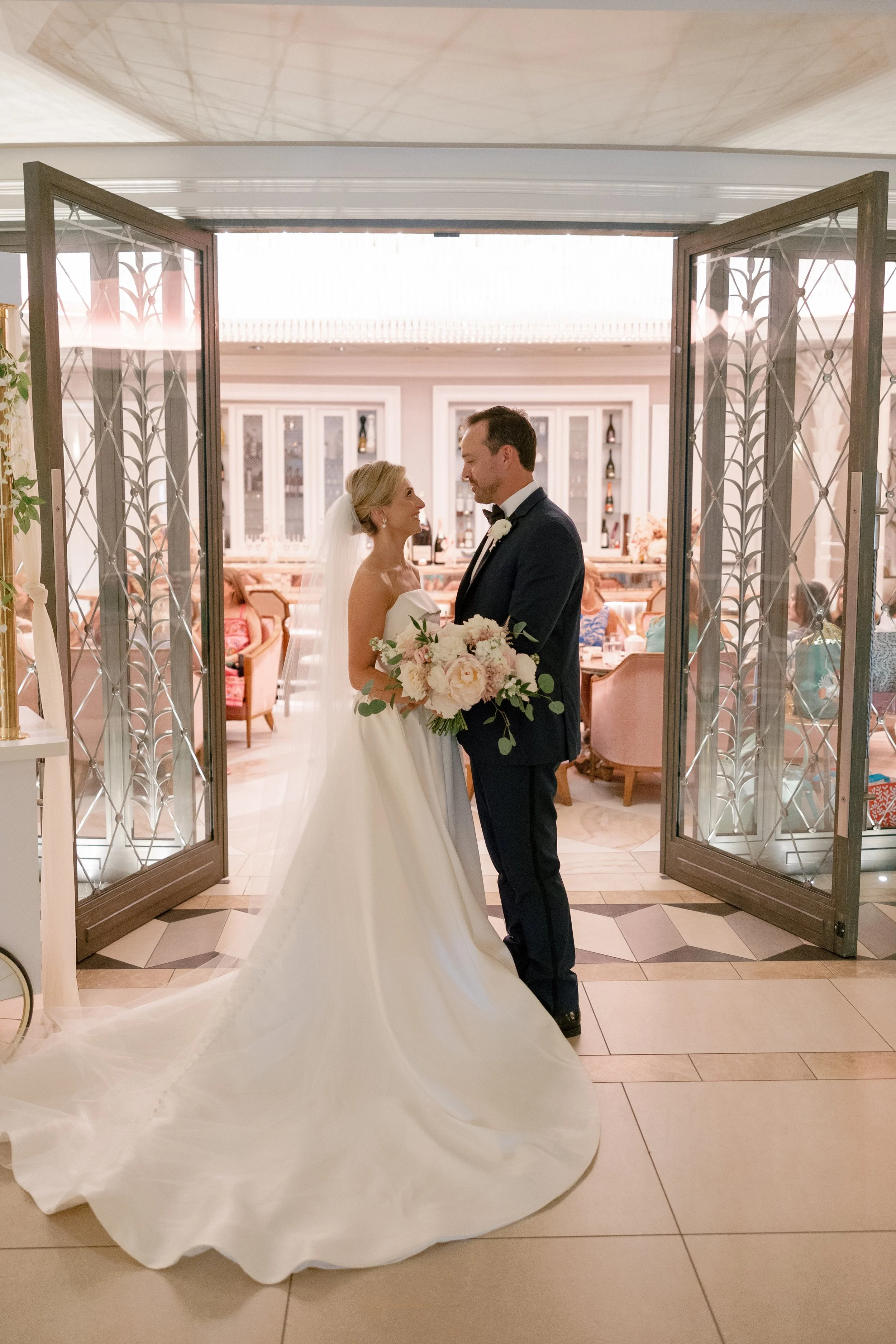 A bride and groom standing together at their wedding, facing each other and smiling. The bride is holding a large bouquet of flowers, and they are standing in front of an open set of decorative glass doors leading into a beautifully decorated reception area.
