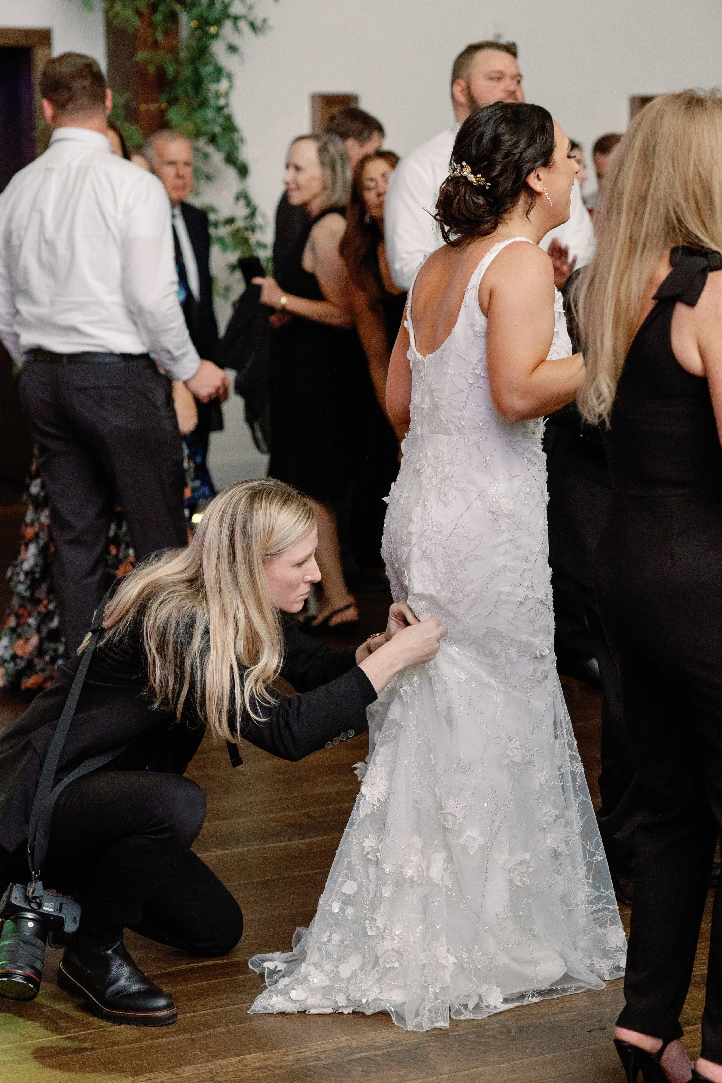 A bride in a white lace wedding dress is having the train adjusted by a woman crouching down at a wedding reception, with other guests mingling in the background.