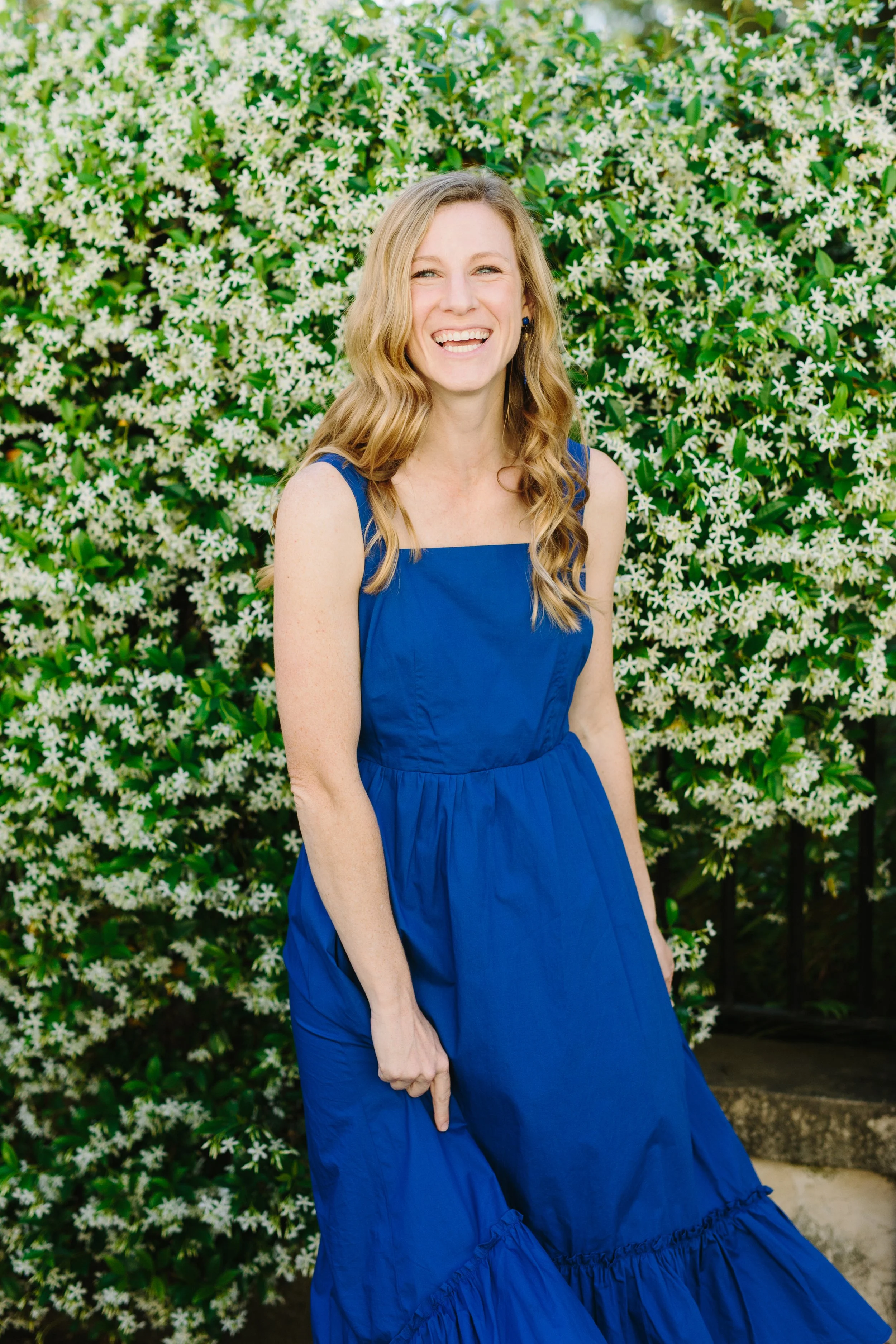 A woman with long blonde hair in a blue dress standing in front of a bush with white flowers, smiling.