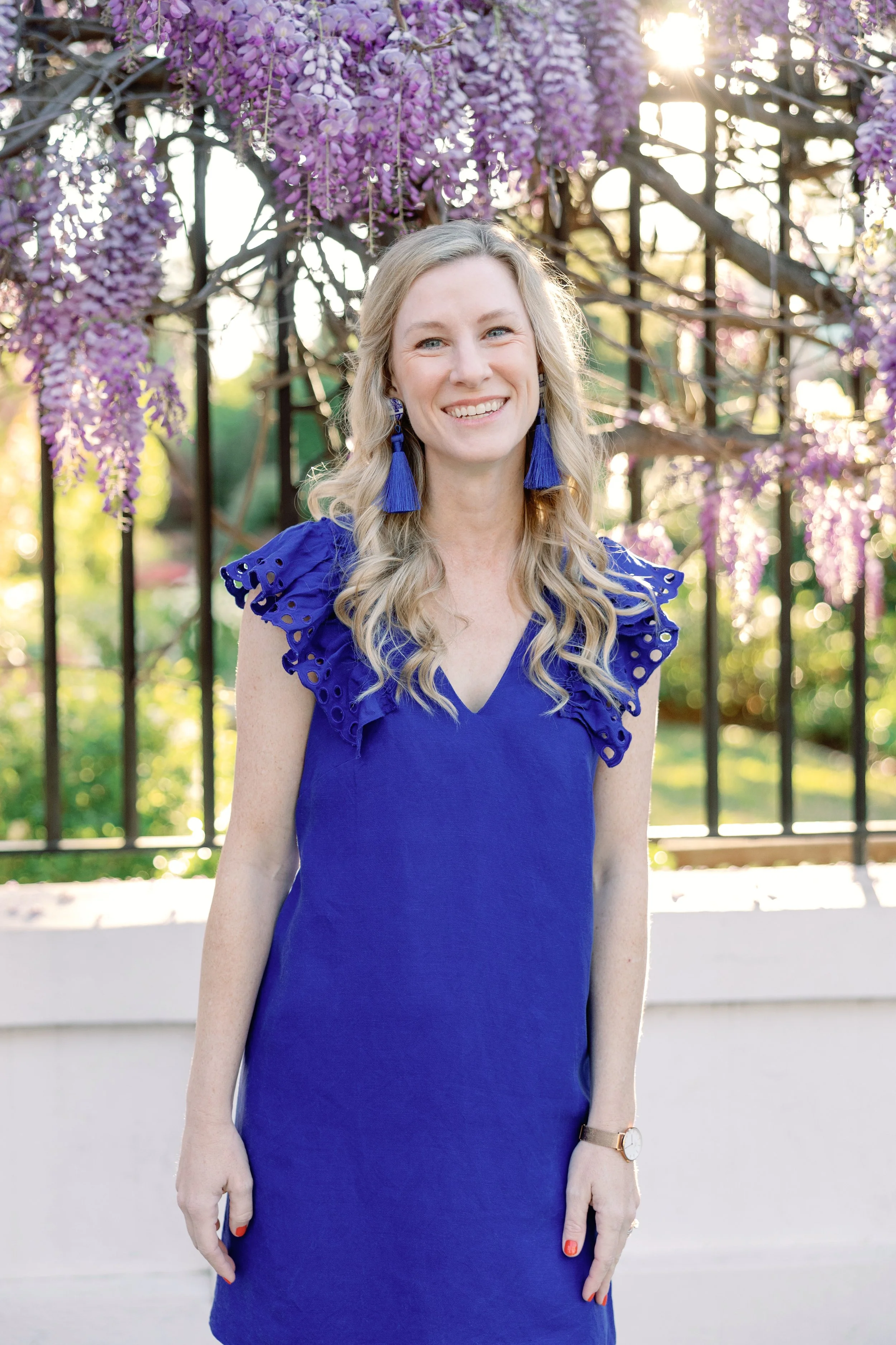 A woman with long, wavy blonde hair smiling in front of purple wisteria flowers hanging from a trellis during sunlight