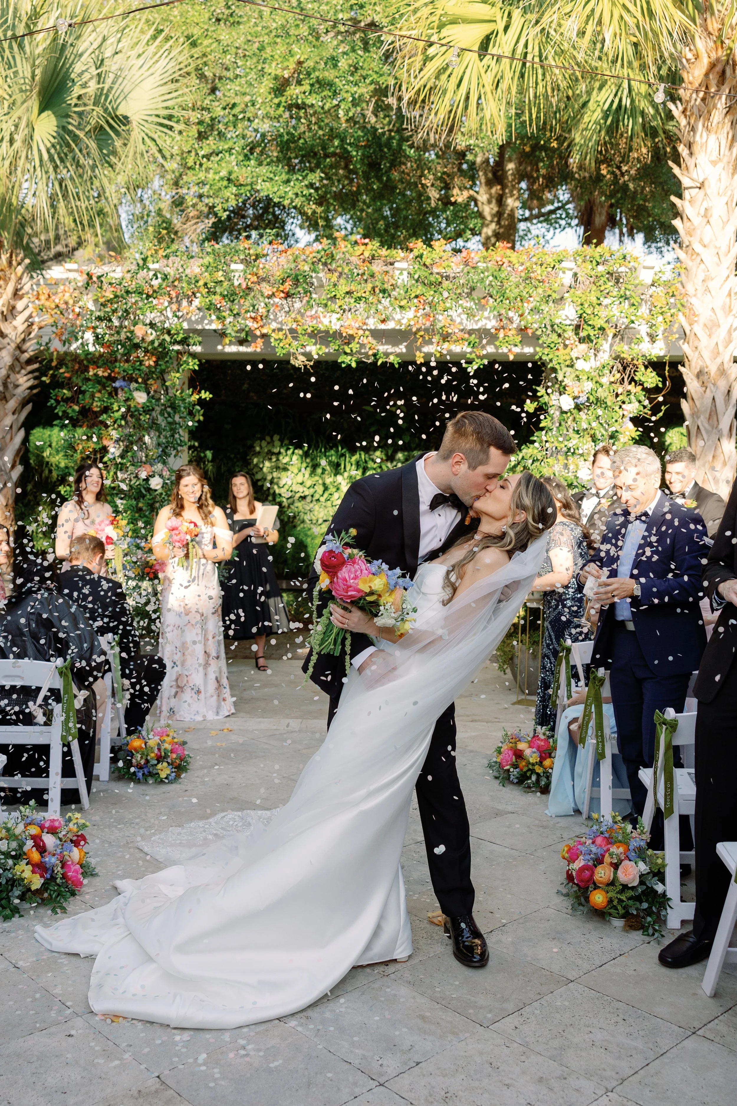 A newlywed couple sharing a kiss under a floral arch at their outdoor wedding ceremony, surrounded by guests and colorful flower arrangements.