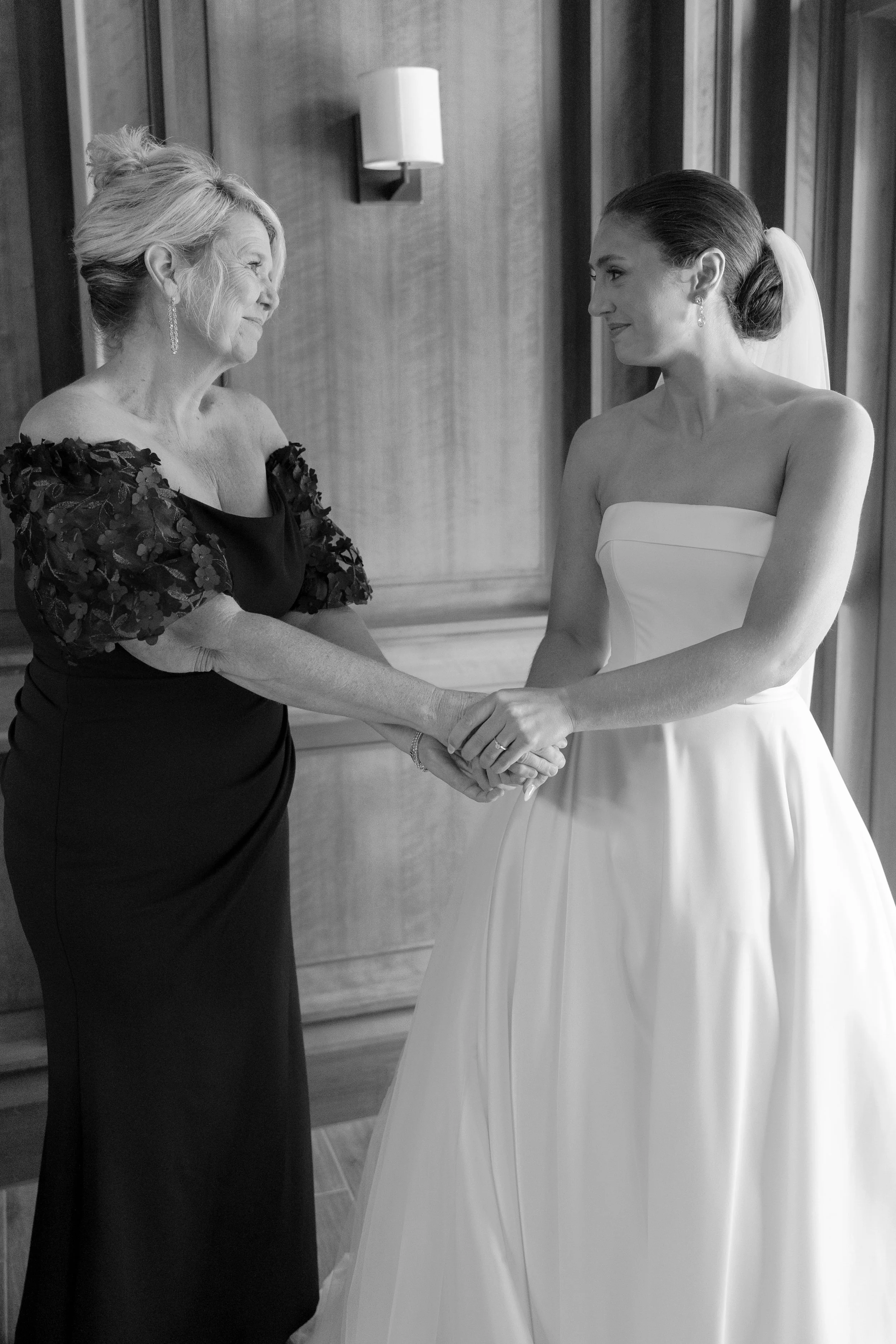 A bride and an older woman holding hands and smiling at each other, inside a room with wooden walls and a wall-mounted light fixture.