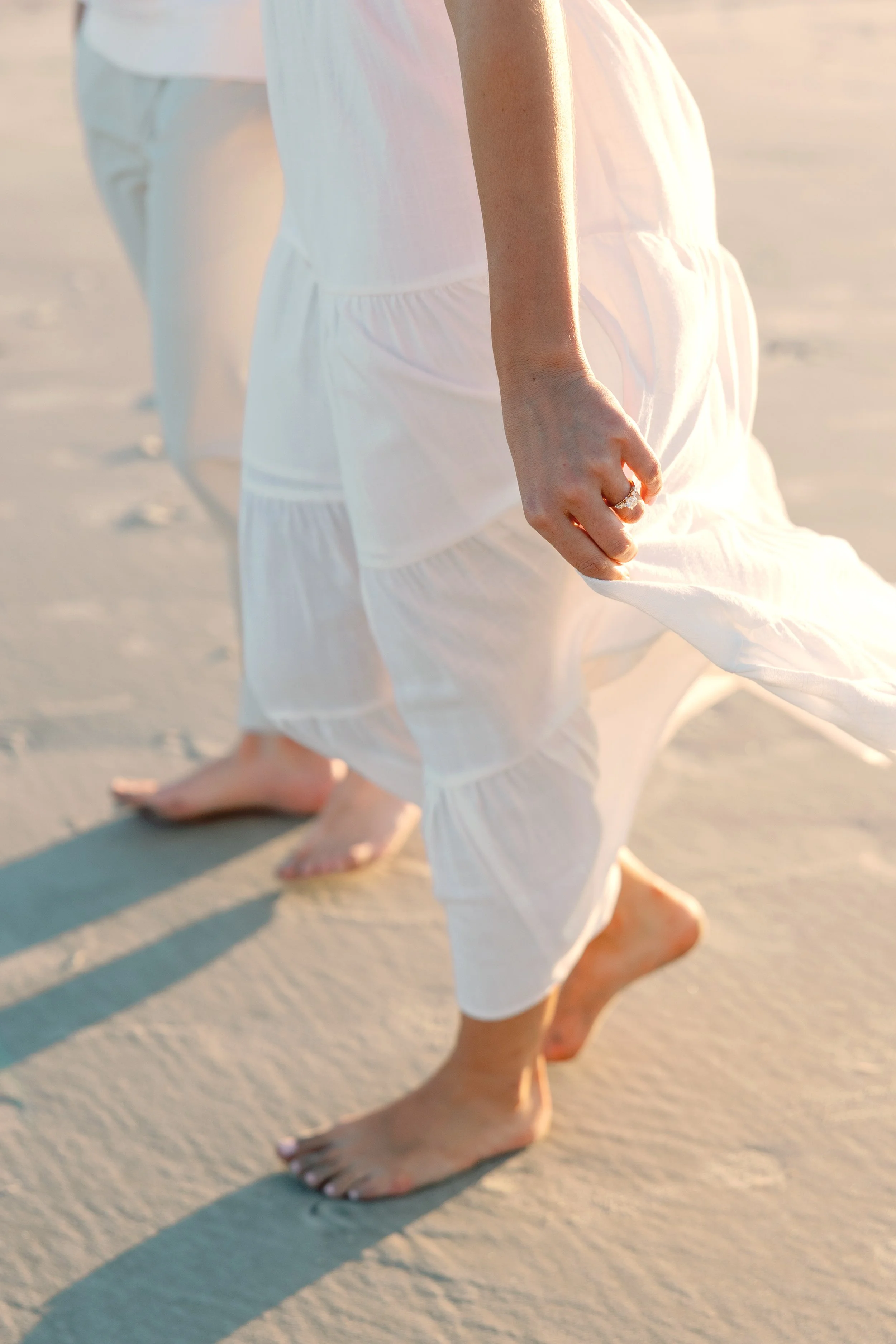 Close-up of two people walking barefoot on a sandy beach, dressed in flowing white clothing, with one person holding their dress.
