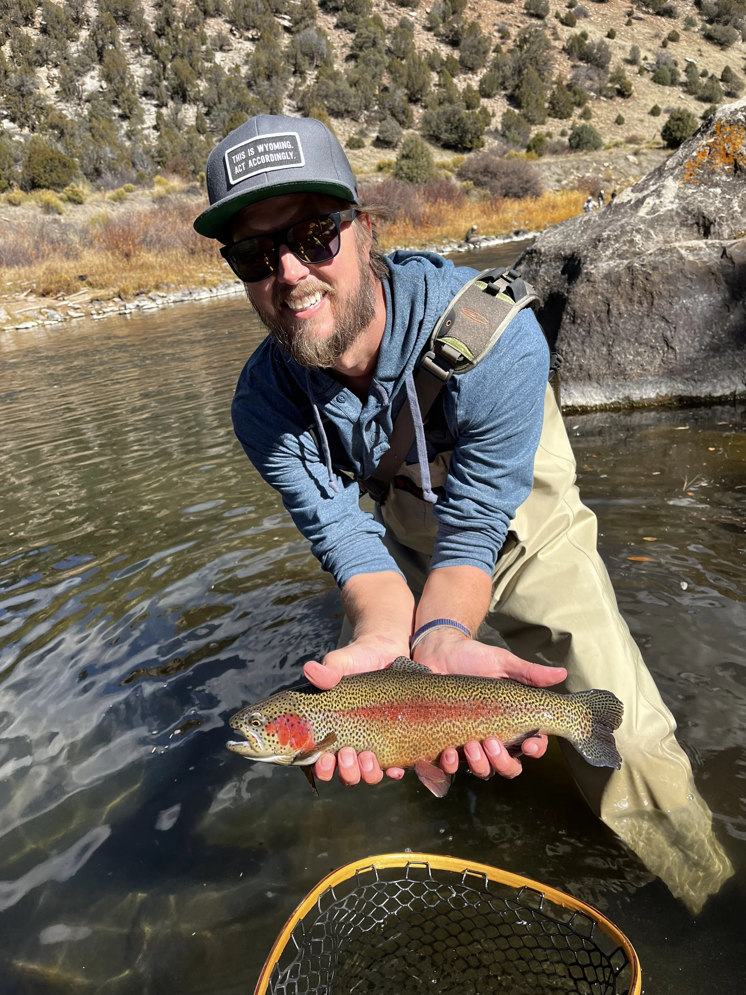 A man wearing sunglasses, a hat, and outdoor clothing is kneeling in a river, holding a colorful rainbow trout with both hands.