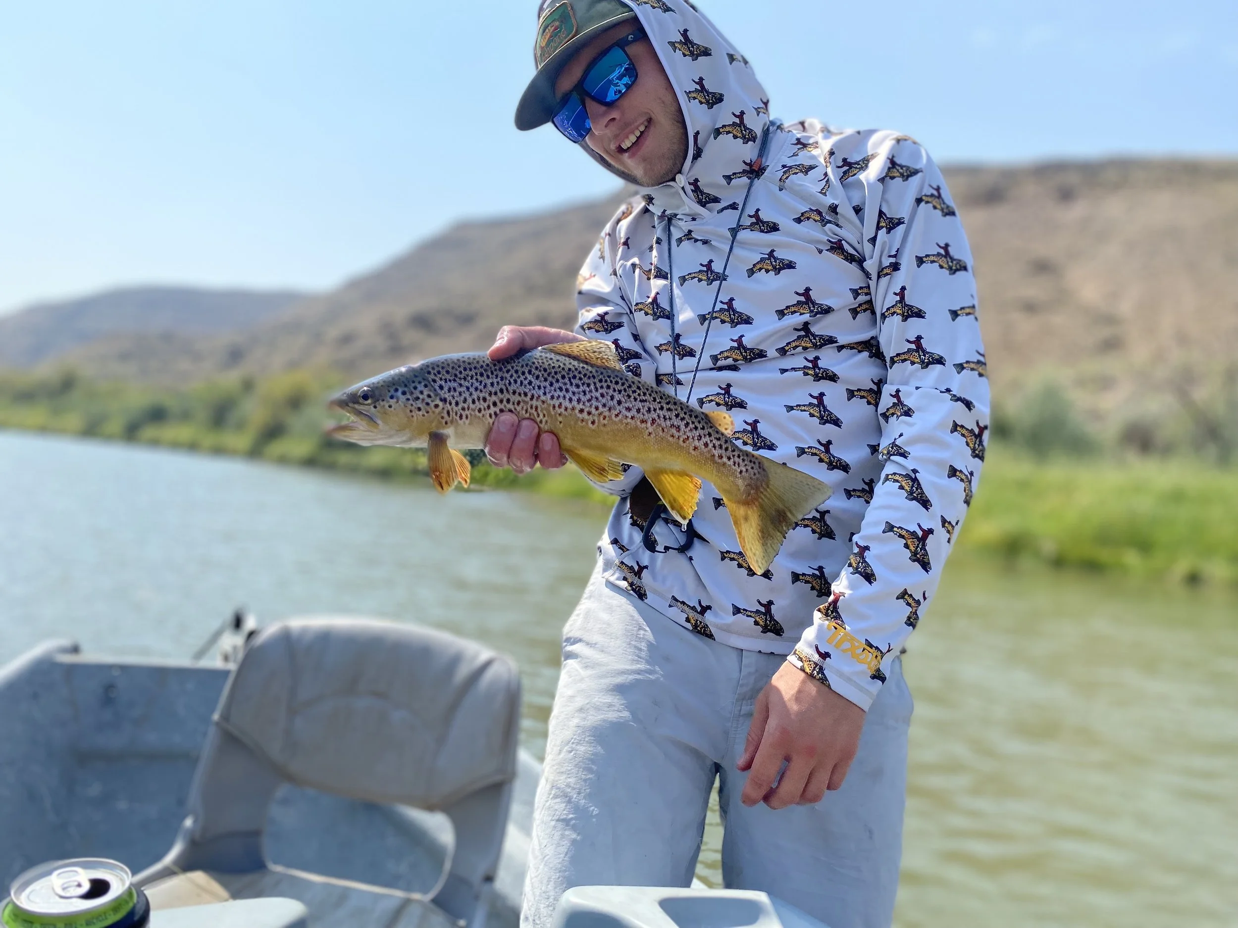A man wearing sunglasses, a cap, and a white jacket with a dinosaur pattern is holding a fish on a boat with mountains and water in the background.