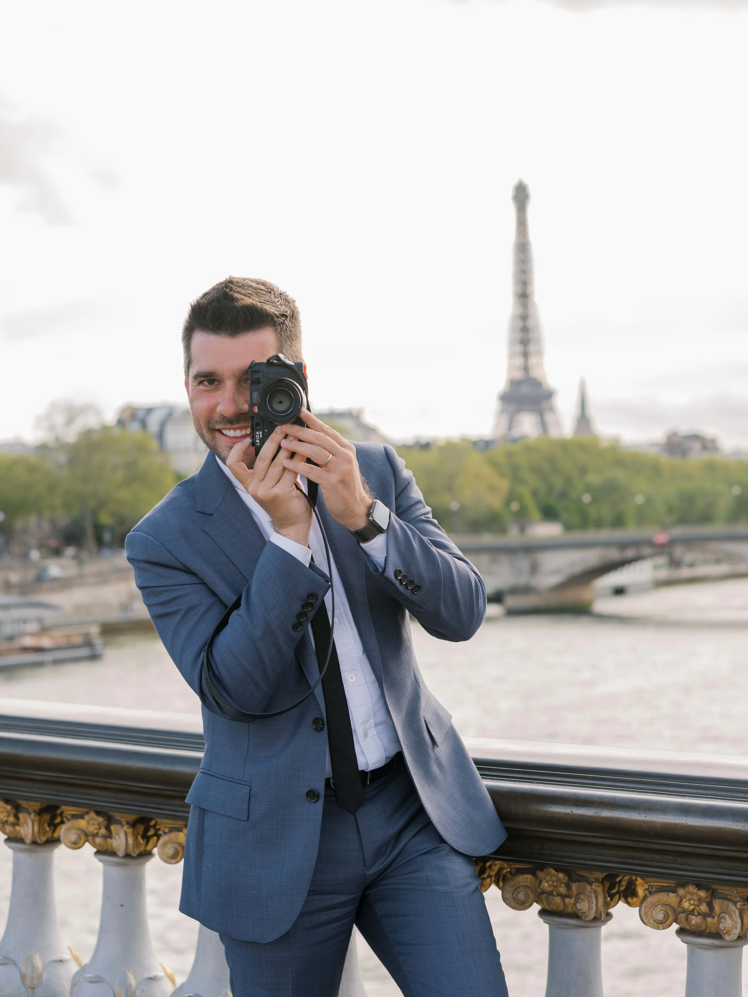 A man in a blue suit taking a photo with a camera near the Seine River with the Eiffel Tower in the background in Paris.