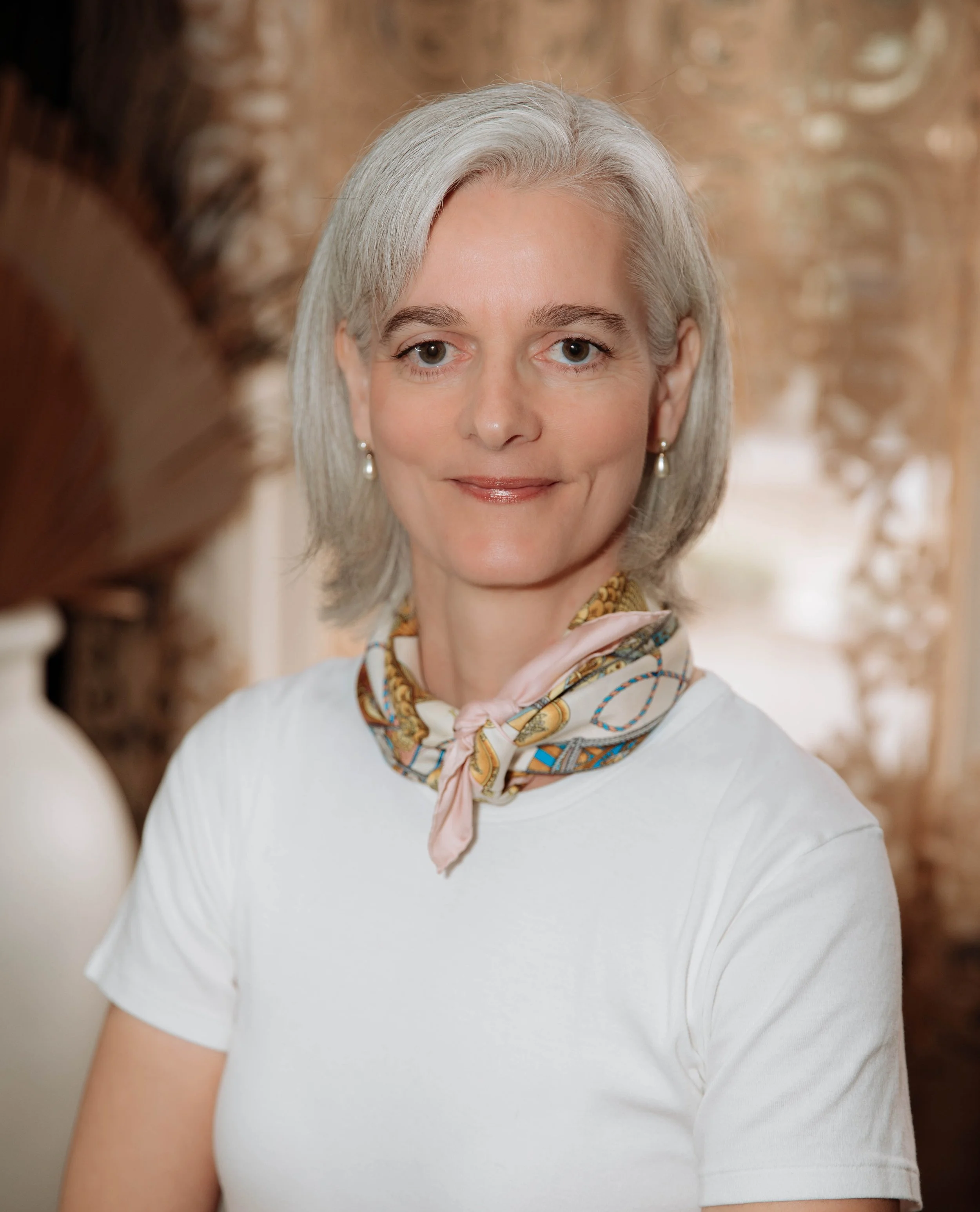 A middle-aged woman with silver hair, wearing a white shirt and a patterned silk scarf around her neck, smiling gently, standing indoors with warm, ornate decor in the background.