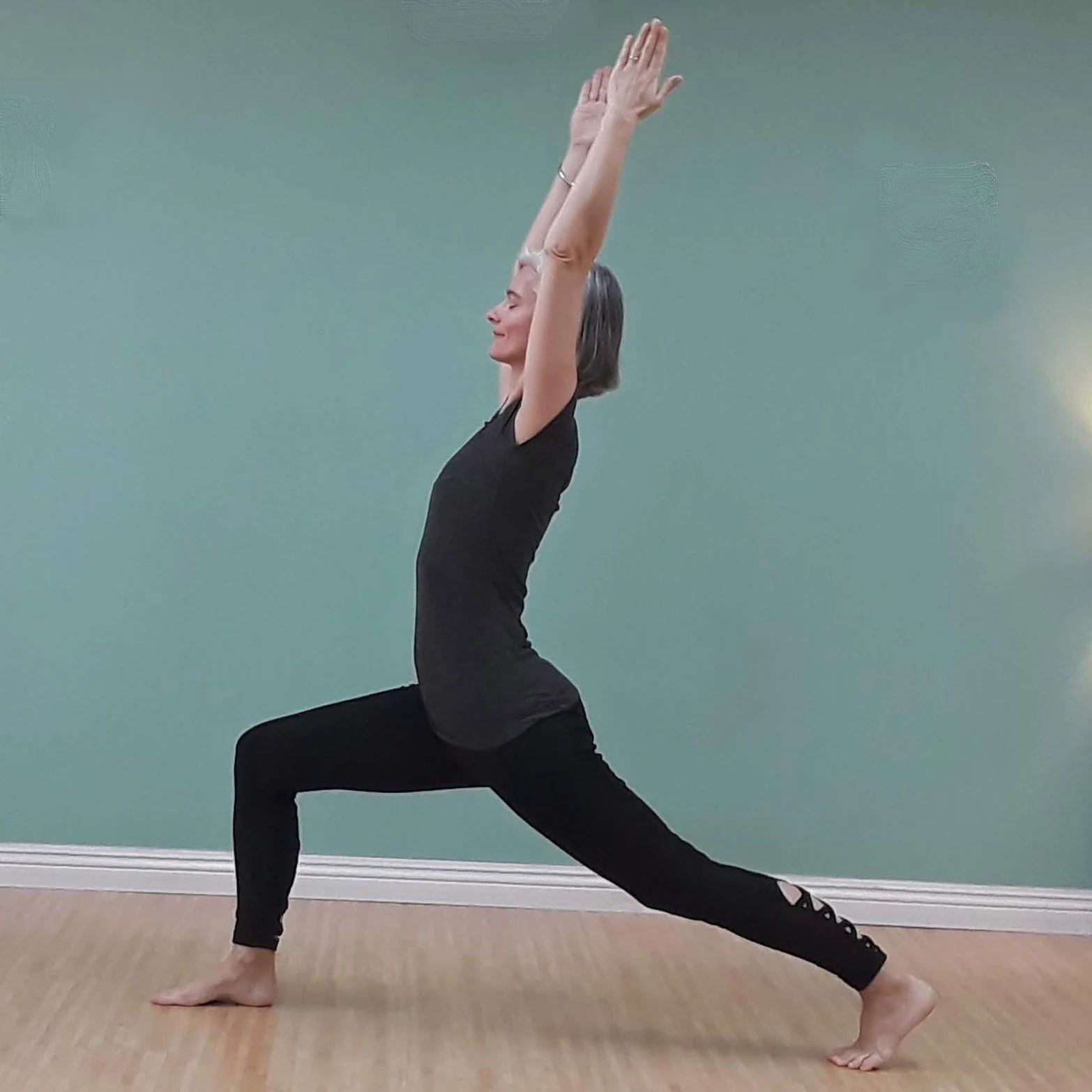 A woman with gray hair practicing yoga in a lunge pose with arms raised overhead indoors against a green wall.