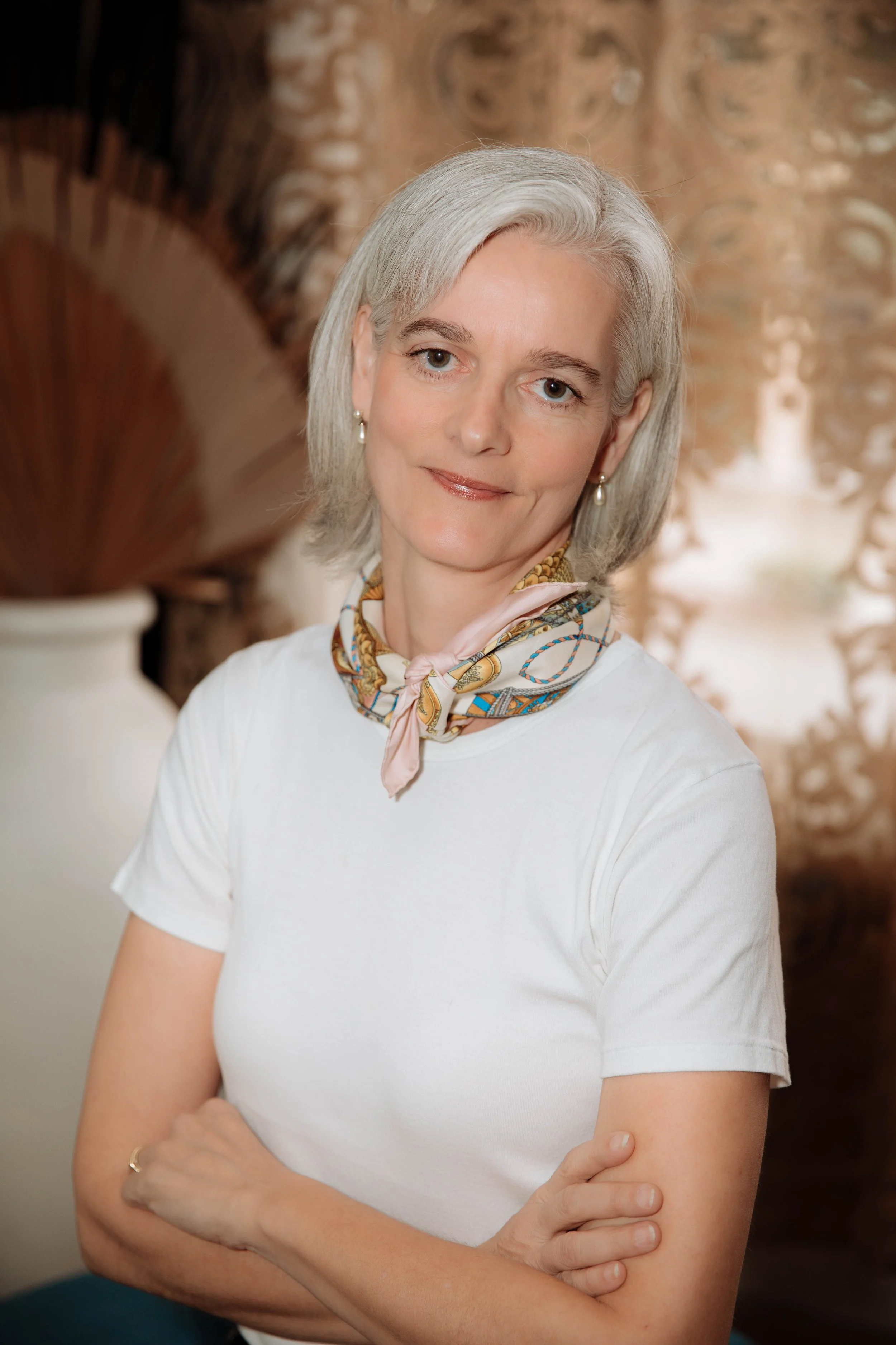 A woman with gray hair and light skin, smiling softly with arms crossed, wearing a white t-shirt and a colorful patterned scarf around her neck, standing indoors with a blurred decorative background.