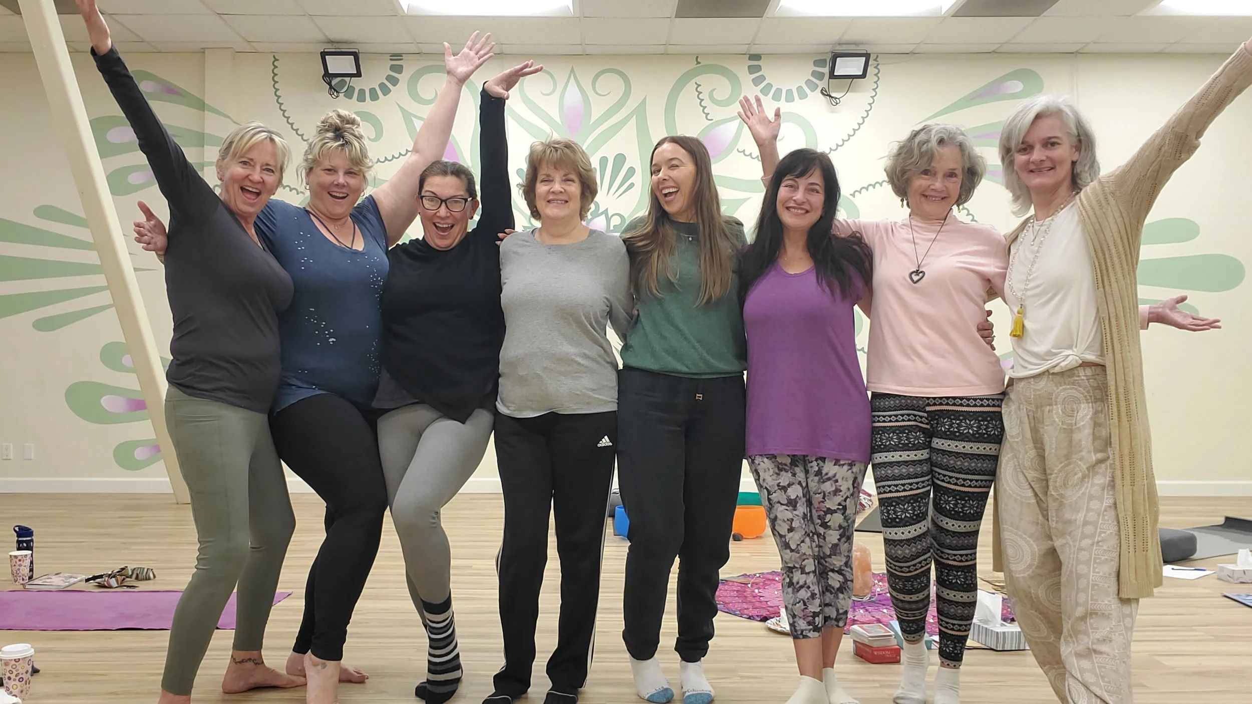Group of eight women standing closely together indoors, smiling and raising their arms in celebration, with some sitting or kneeling, colorful mural on the wall behind them, yoga mats and personal items on the floor.