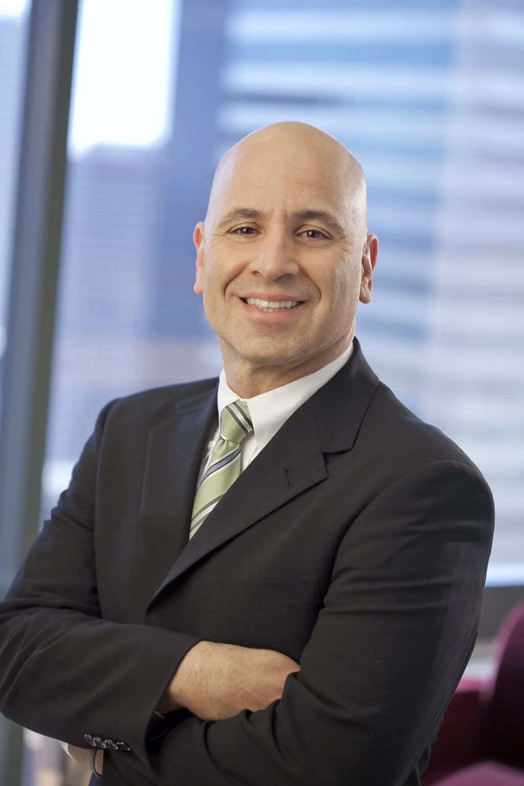 Smiling man with a clean shaved face and head, wearing a white shirt with a balck blazer and a gray tie with his hands across his chest standing against the backdrop of highrises