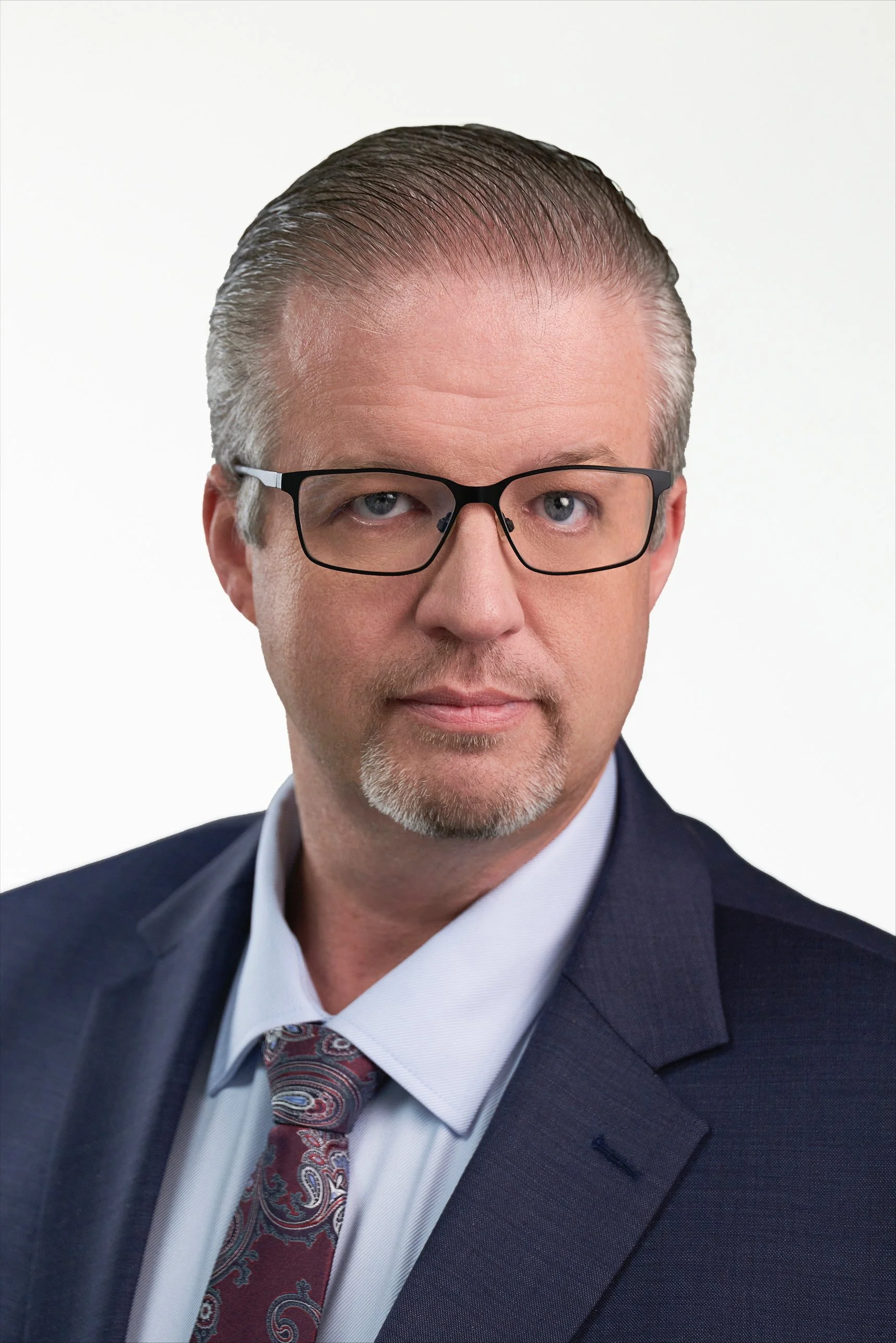 Man wearing a black framed spectacles, a light grey brown french beard, hair combed back, wearing a white shirt with black blazer and a silk tie against a white background
