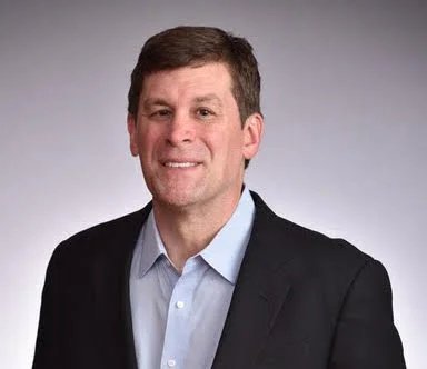 Smiling man with a clean shaved face, dark hair combed back, wearing a white shirt with a black blazer
