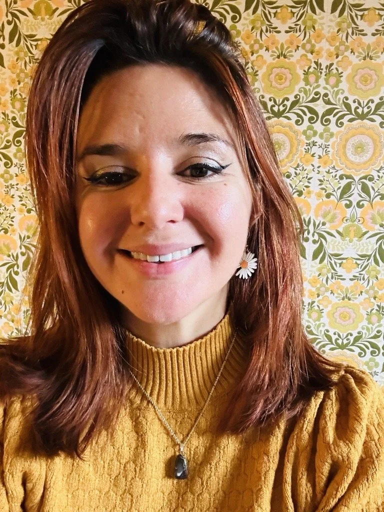 A smiling woman with reddish-brown hair, wearing earrings, a necklace with a heart pendant, and a yellow turtle neck sweater, against a wall with yellow wallpaper with gray patterns