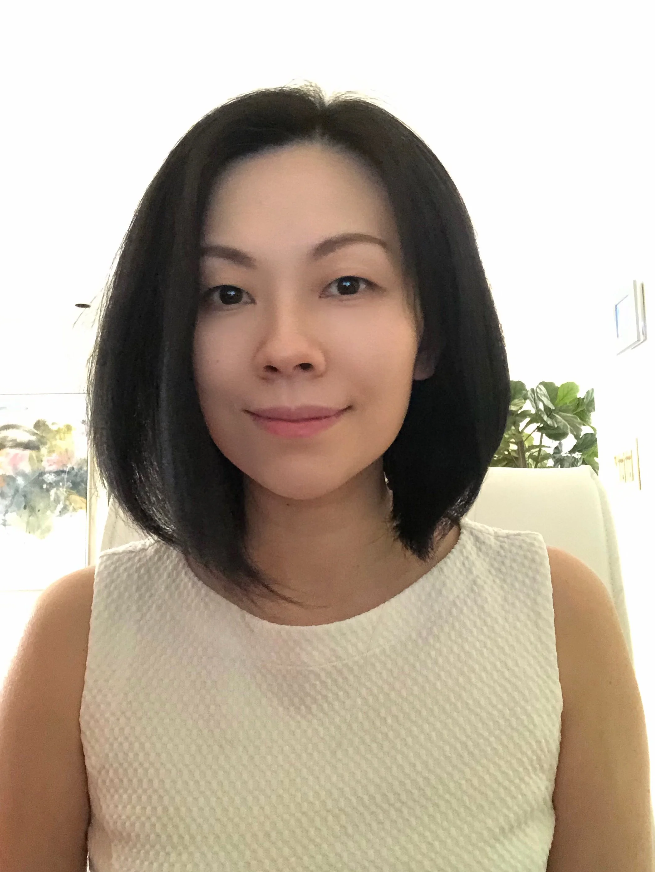 Smiling woman with shoulder length, straight black hair,  wearing a white cotton sleeveless top, against a background of trees and sky afar