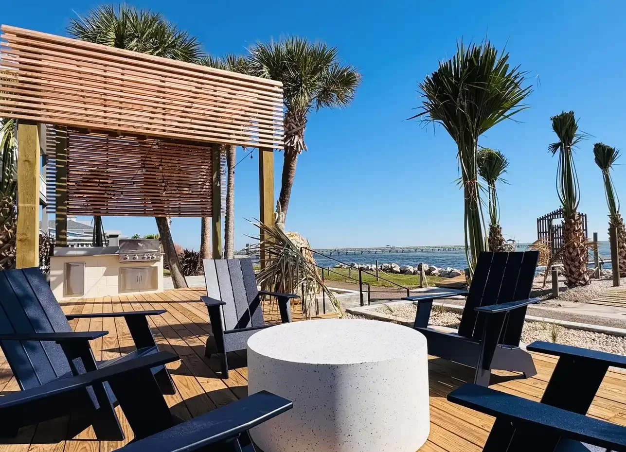 Waterfront deck at Skiff House with Adirondack chairs, fire table, outdoor grill, and scenic bay views framed by palm trees.