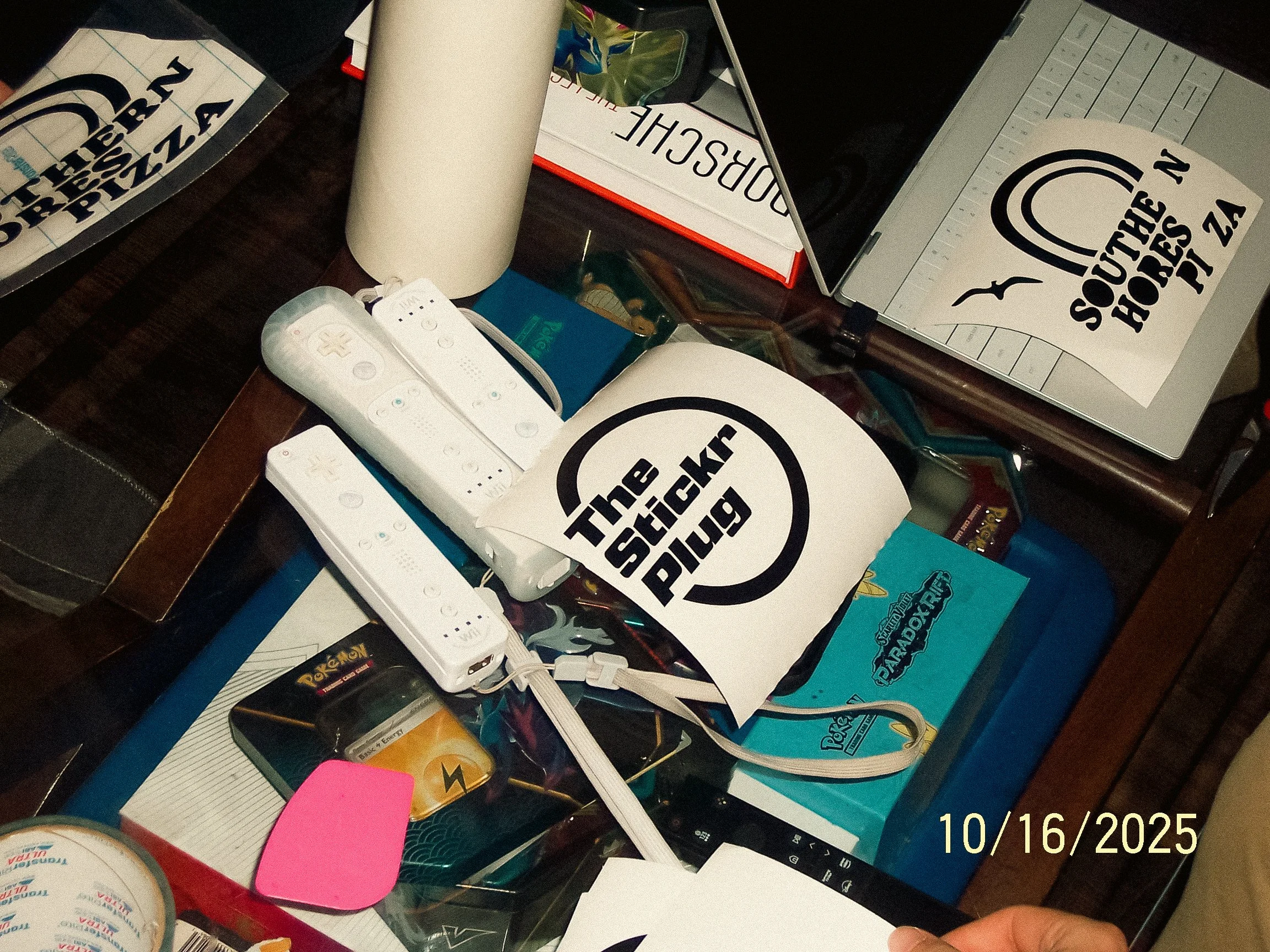 Photo of a cluttered table with various items, including a white TV remote, numerous stickers and packaging, sheets with logos and text, a white mug, and miscellaneous papers and objects.