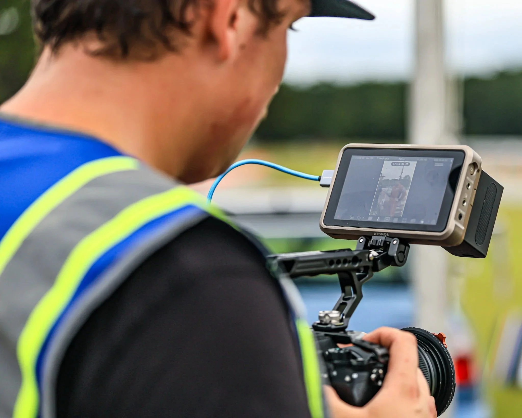 A person wearing a high-visibility vest is looking at a camera or recording device mounted on a tripod outdoors.