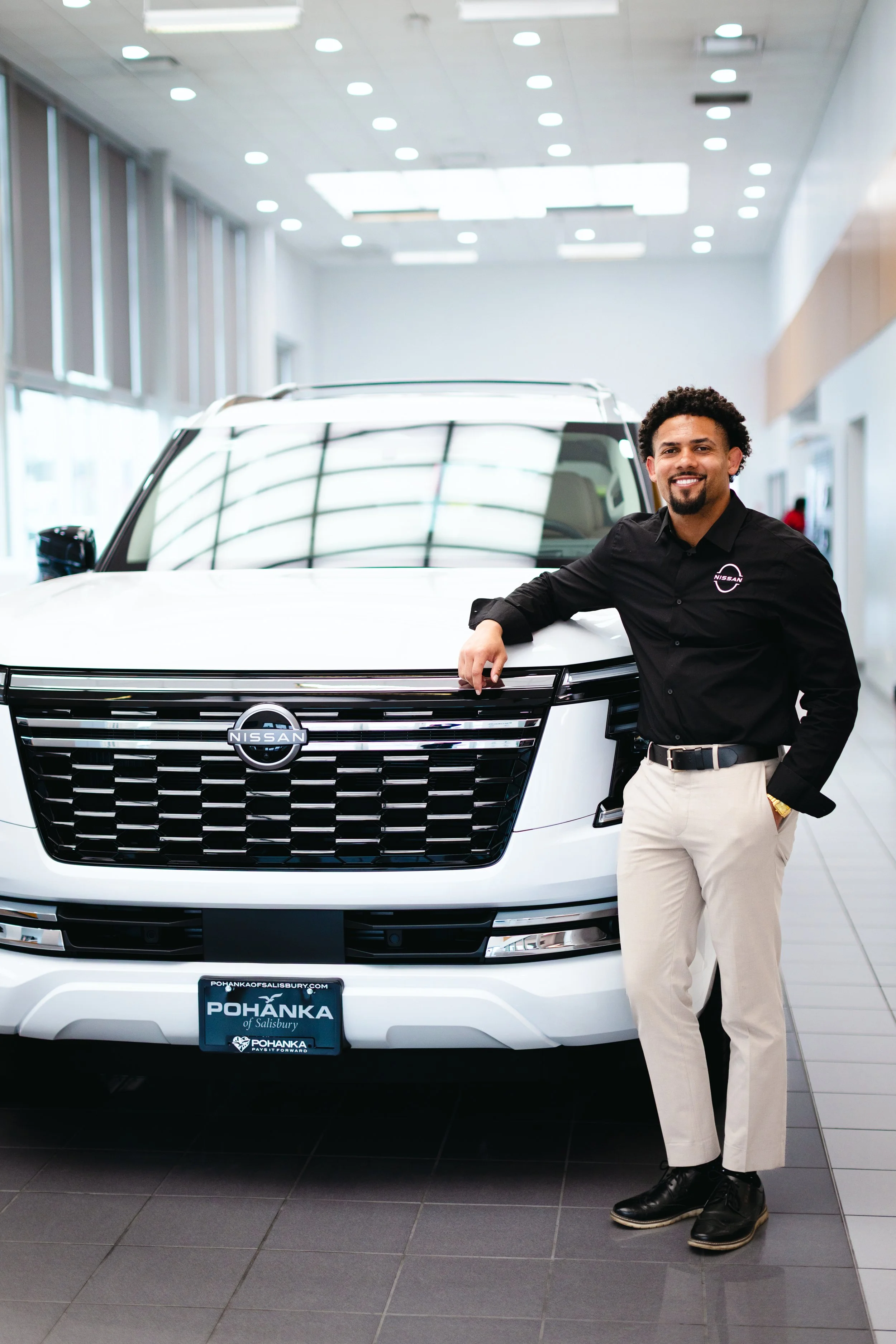 A smiling man in black shirt and beige pants standing next to a white Nissan SUV inside a car dealership.