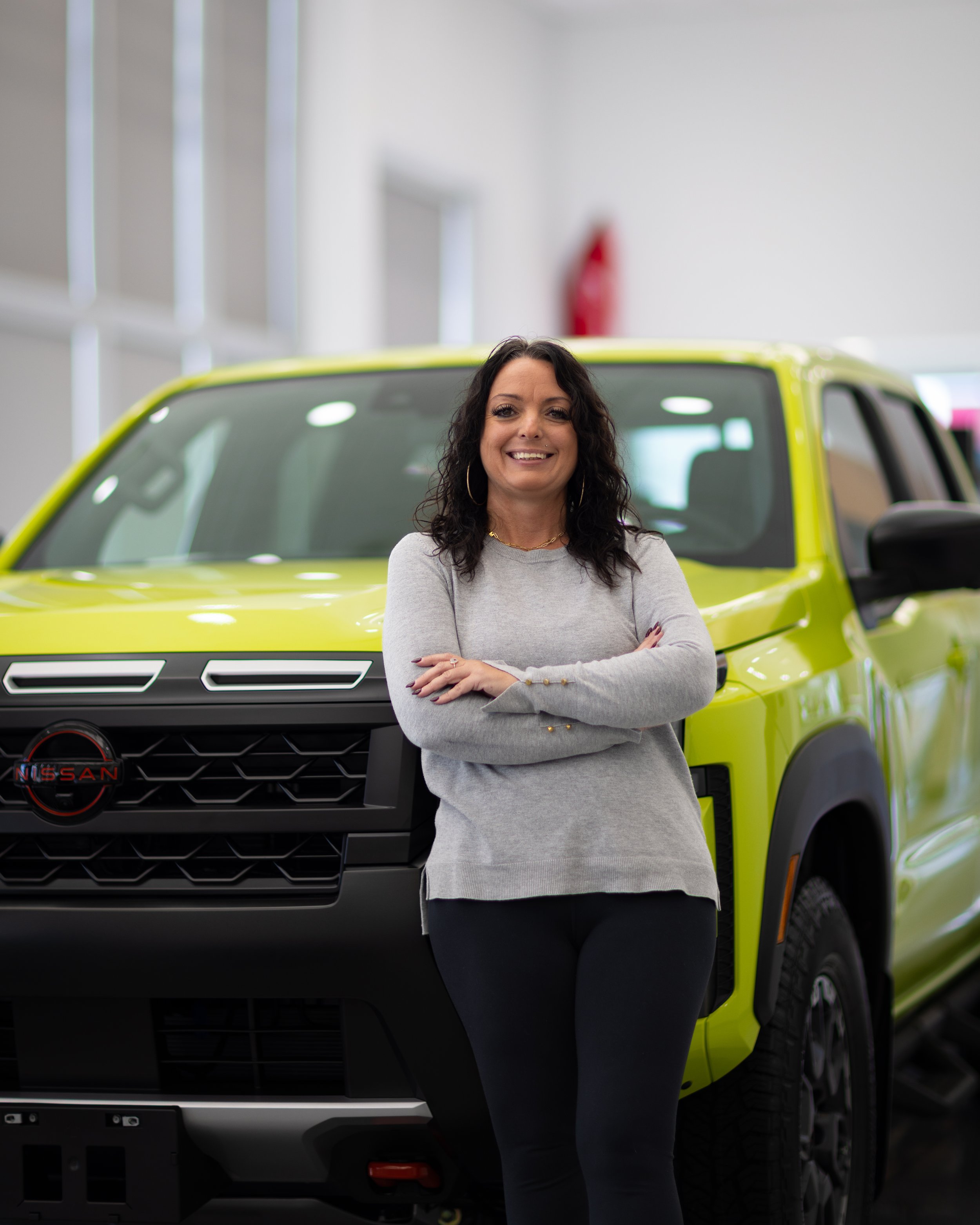 A woman with dark curly hair, gold hoop earrings, and a light gray long-sleeve shirt standing in front of a bright yellow-green Nissan SUV inside a showroom, smiling with crossed arms.