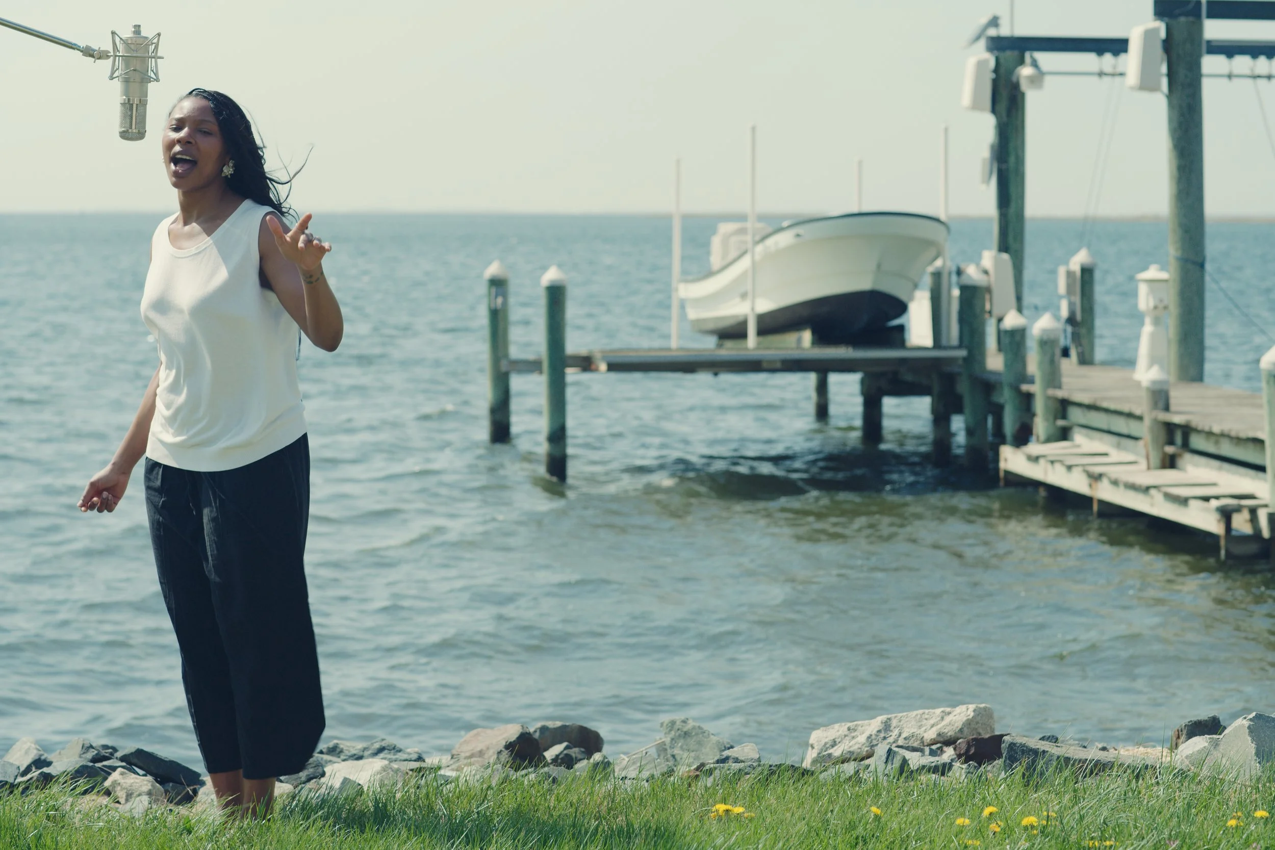 A woman standing by the water near a dock and boat, singing into a microphone on a stand.