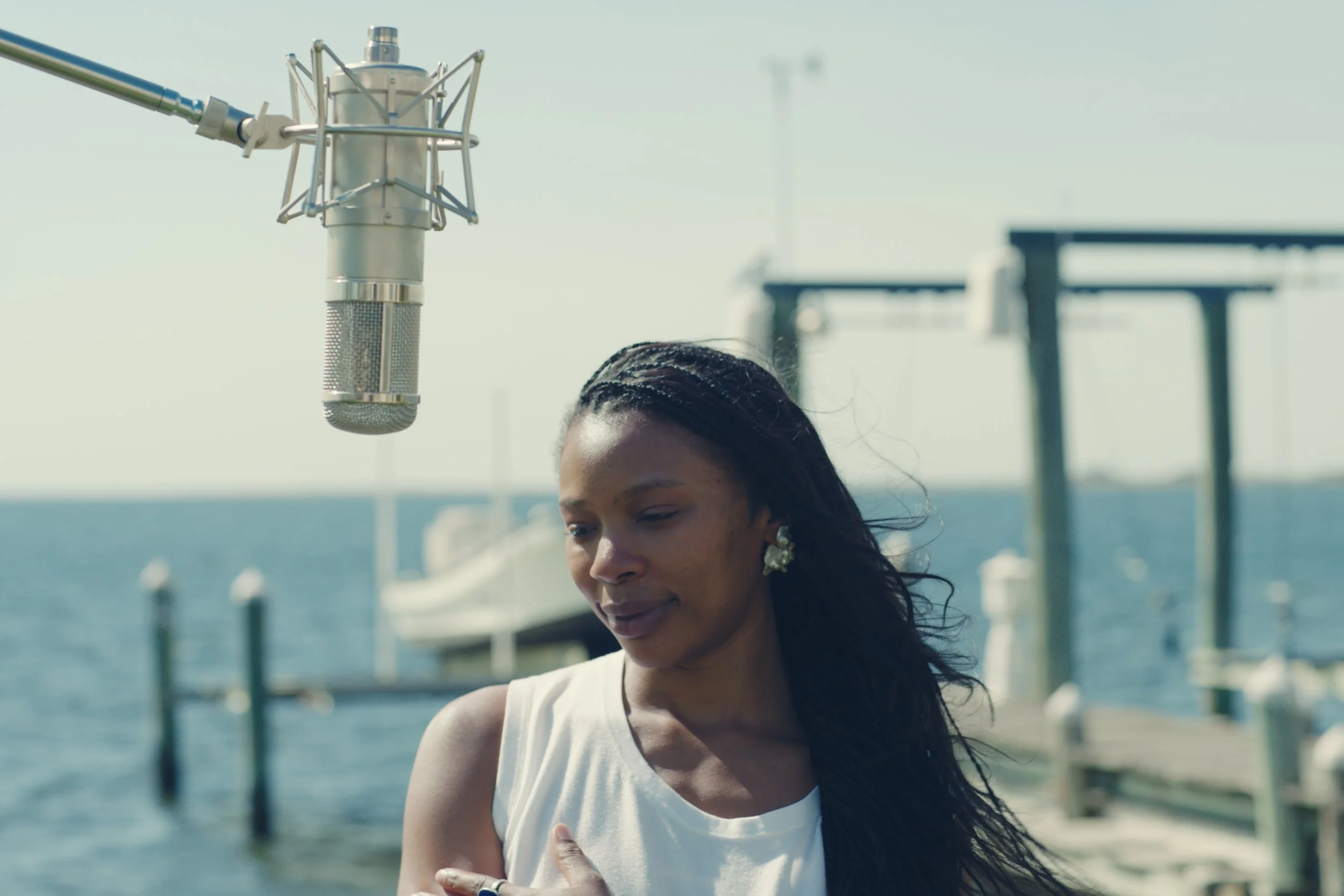 A woman with long dark hair wearing earrings and a white top, standing outdoors near a body of water with a microphone hanging above her.