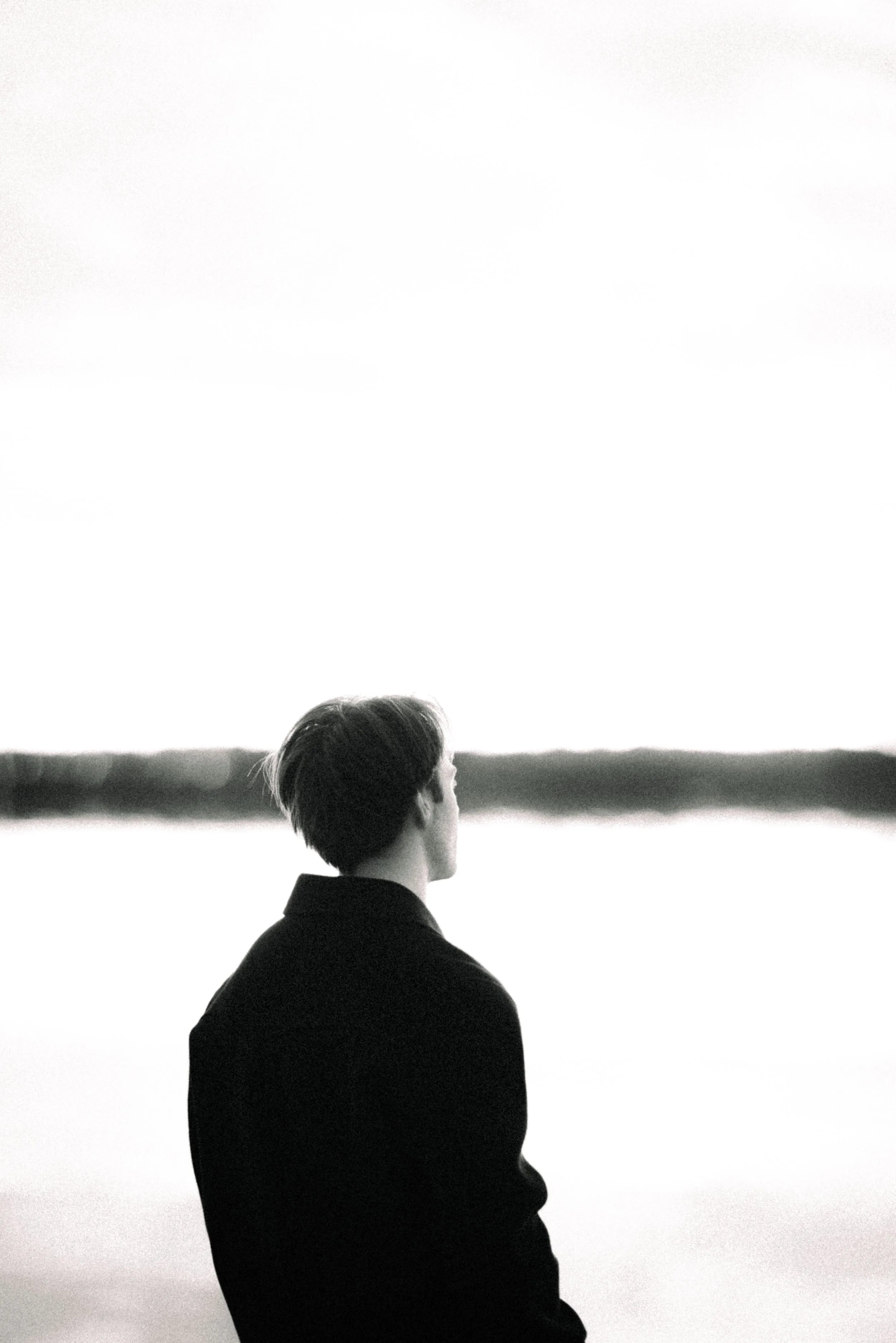 A person with short hair, viewed from behind, sitting outdoors and looking toward a horizon with open space and a body of water or land in the distance, with a calm and contemplative atmosphere.