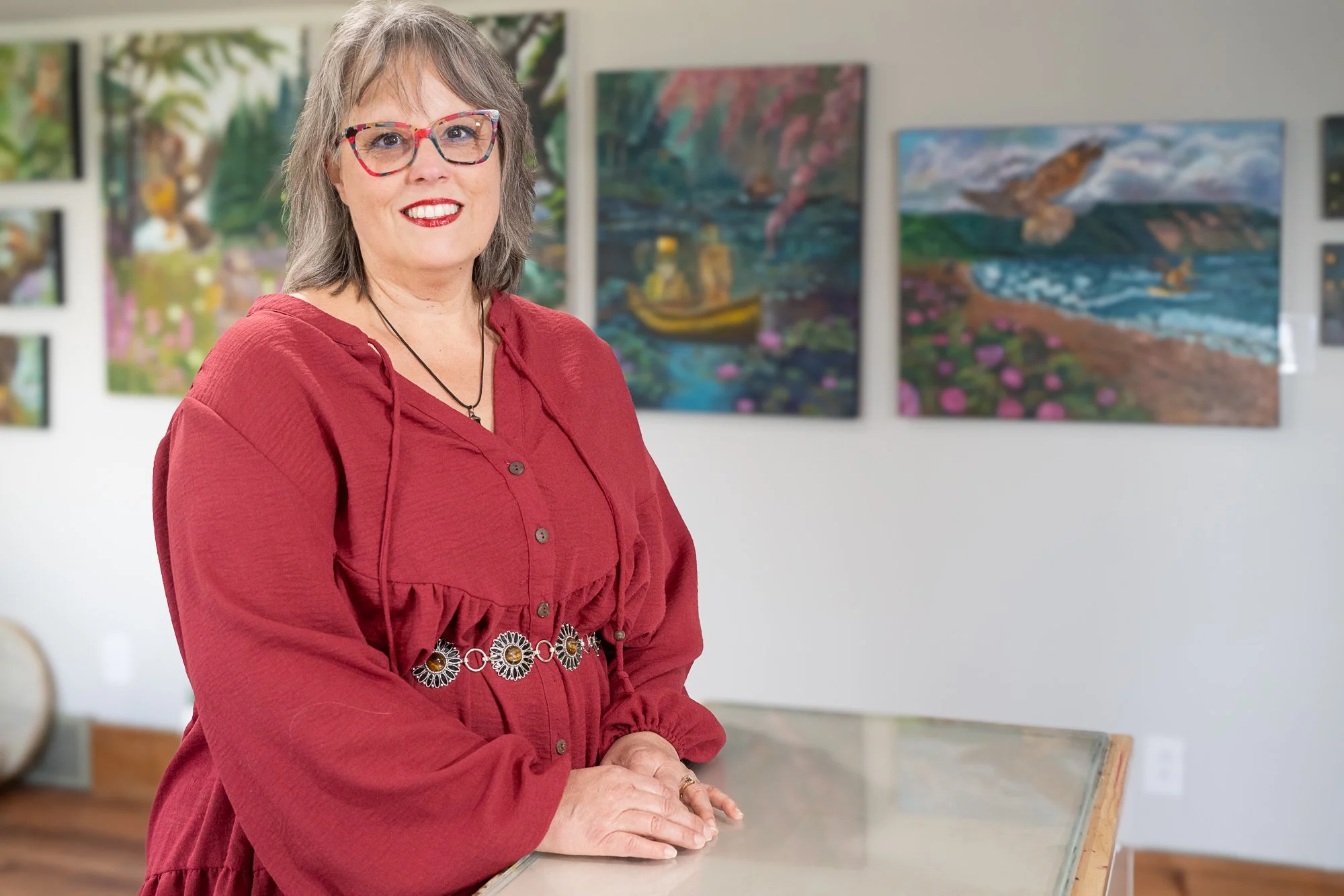 A woman with gray hair and glasses standing in an art gallery, smiling and leaning on a glass display case, with colorful landscape paintings in the background.