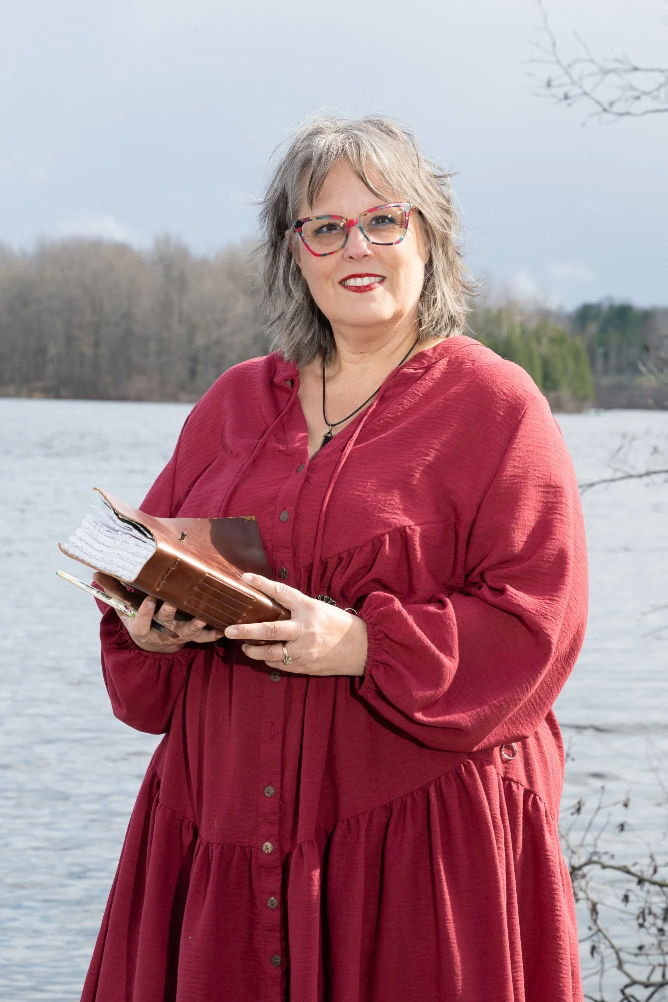 A woman with gray hair, wearing colorful glasses and a red dress, smiling and holding a book, outdoors near a lake with trees in the background.
