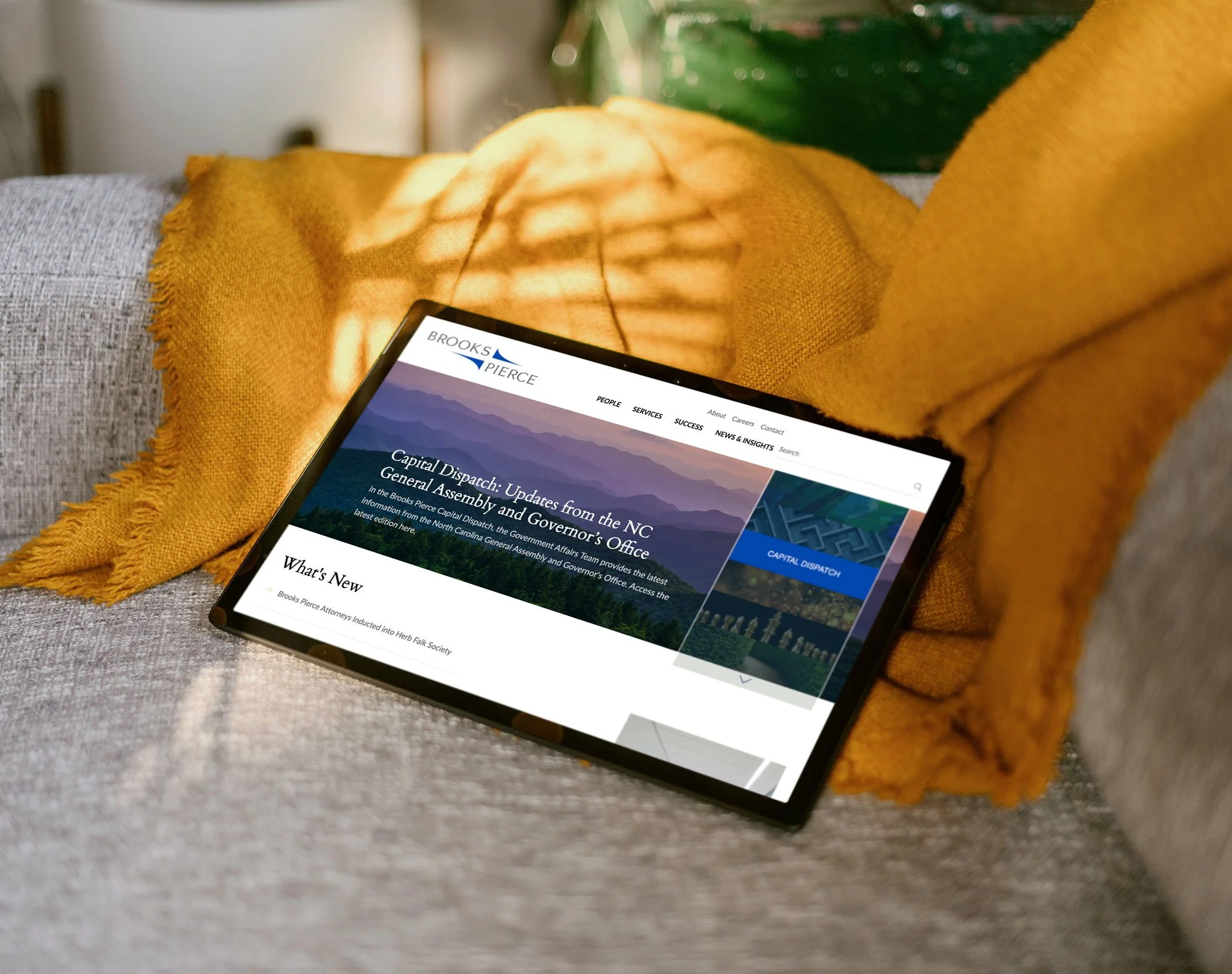 Tablet displaying Brooks Pierce law firm website homepage with a clean blue-toned layout, resting on a couch with warm accent blanket.