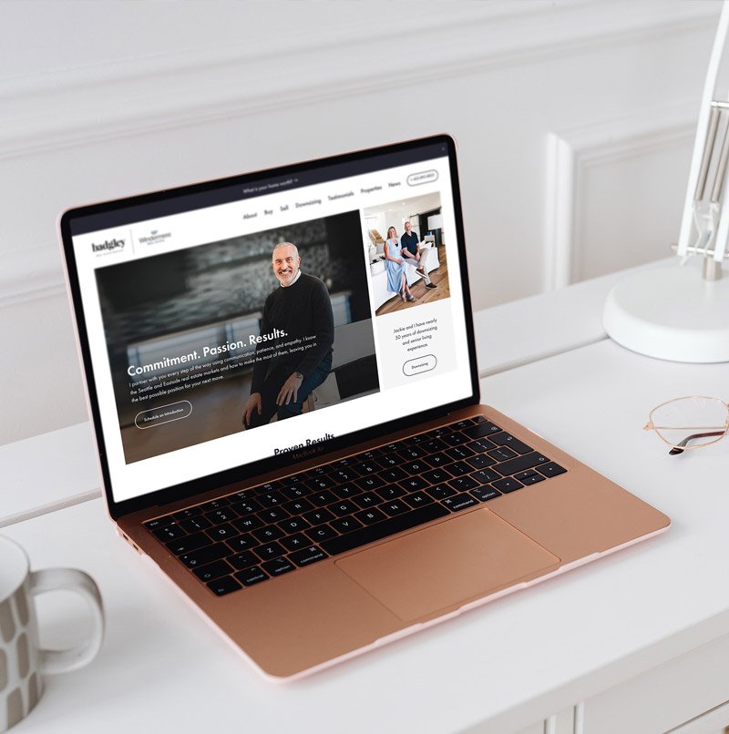 Laptop displaying Badgley Homes real estate website homepage with a clean, modern layout, shown on a minimal desk with coffee mug and glasses.