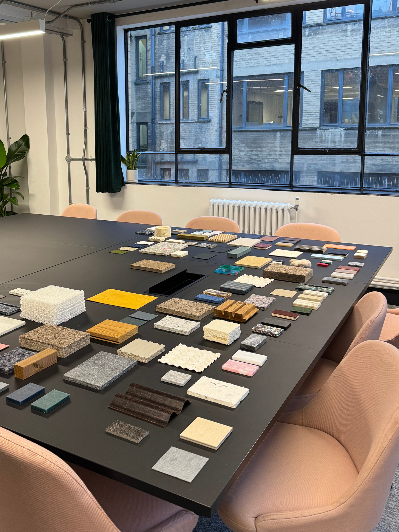An office conference room with a large black table displaying various color and material samples, surrounded by pink upholstered chairs. Large windows with black frames show a brick building outside, and there are green plants and a radiator inside.